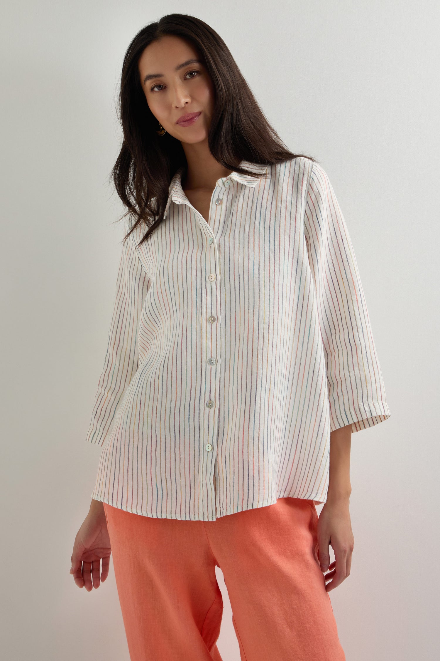 A woman stands against a plain background wearing a White Space Dye Stripe Linen Shirt and coral-colored pants.