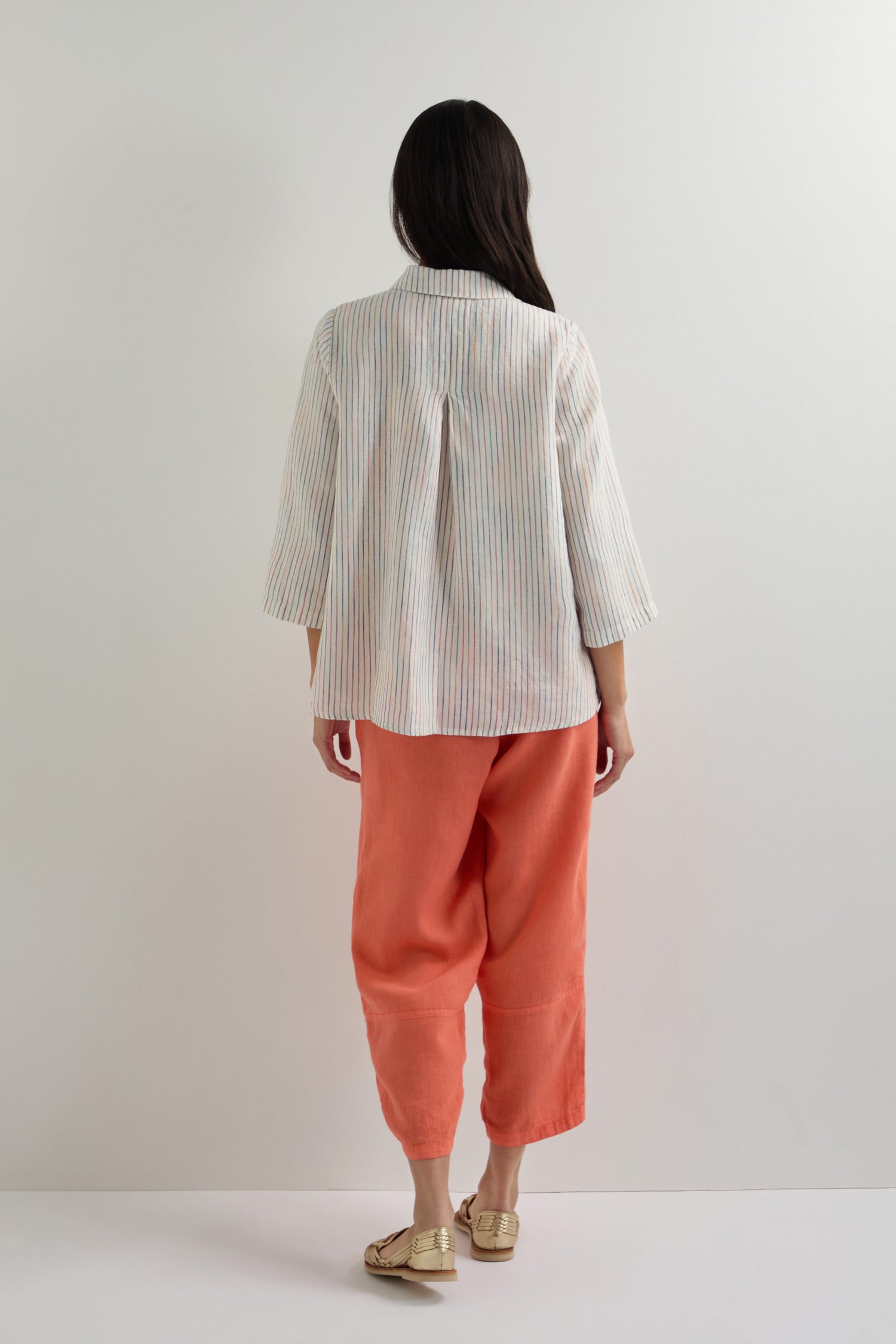A person stands facing a white wall, wearing a loose, classic fit White Space Dye Stripe Linen Shirt, coral pants, and beige shoes.