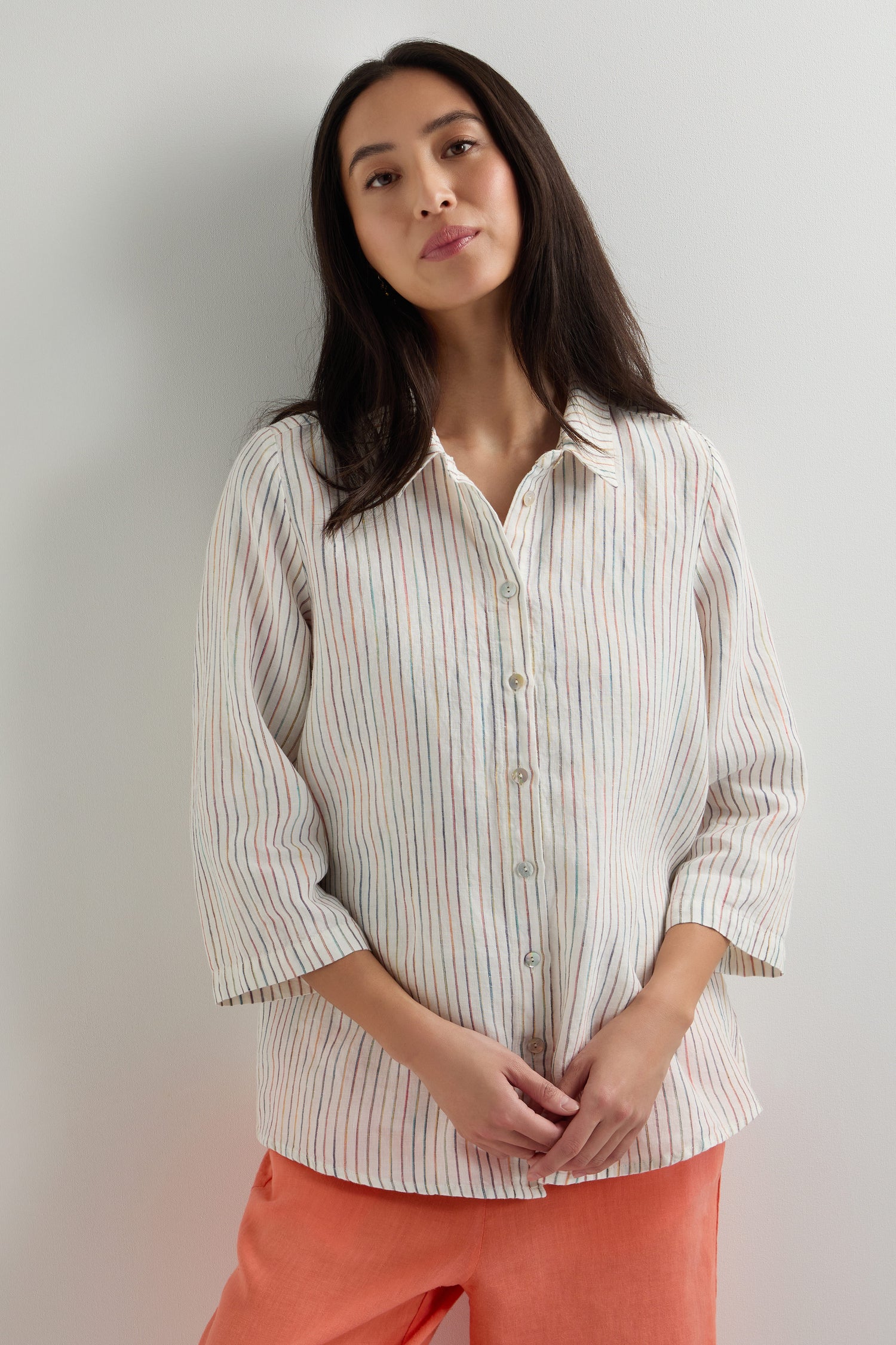 A woman in a classic fit White Space Dye Stripe Linen Shirt with long sleeves and orange pants stands against a plain, light-colored wall.
