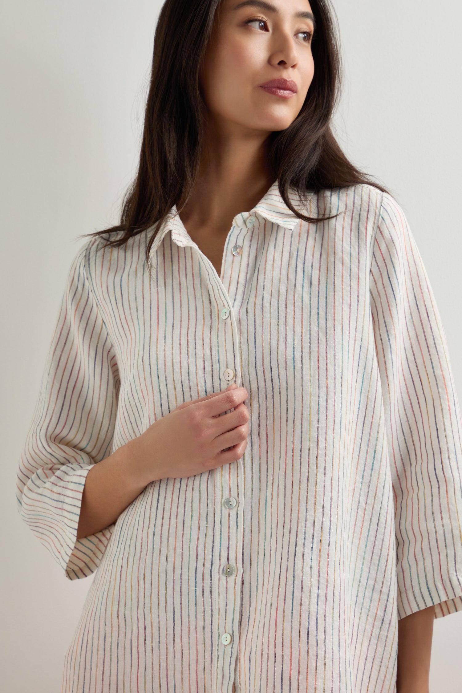 A woman wearing a White Space Dye Stripe Linen Shirt with loose long sleeves and vertical pastel stripes stands against a plain light-colored background.