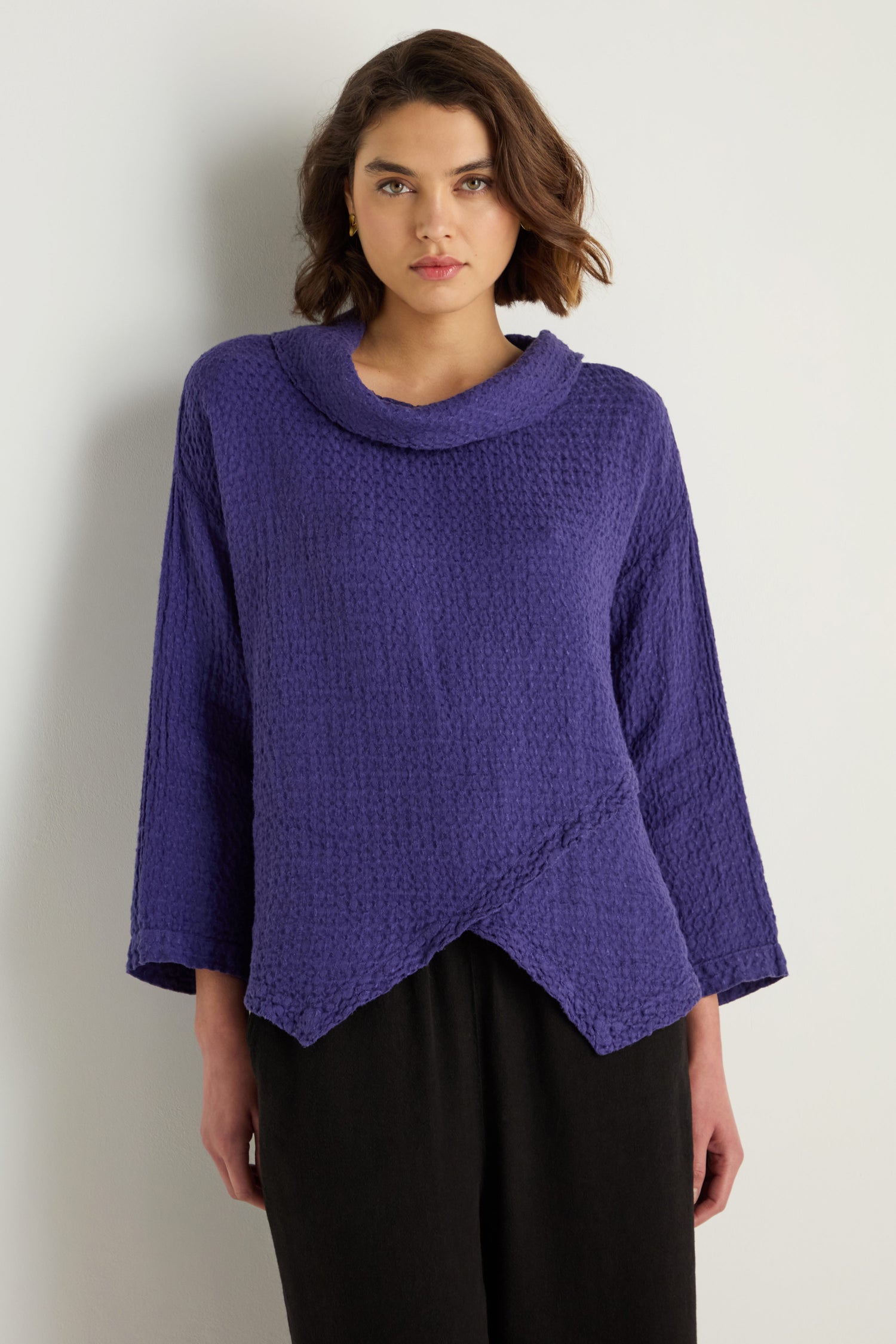 A person stands against a light background wearing the Waffle Linen Boxy Cowl Neck Top in purple, featuring a textured waffle finish, asymmetrical hem, relaxed sleeves, and black pants.