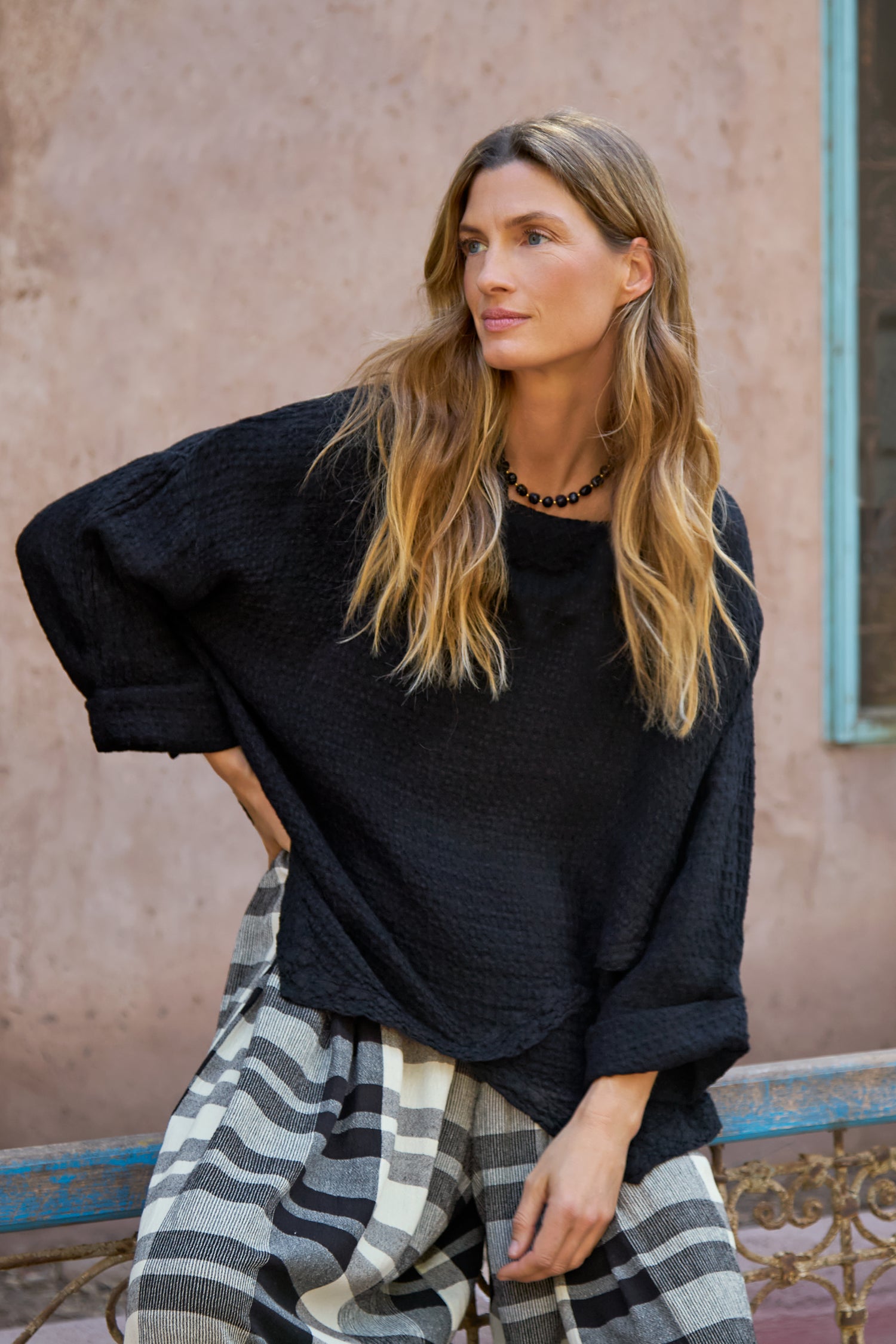 A woman with long, wavy hair wears the Waffle Linen Boxy Cowl Neck Top in black with checkered pants, sitting by a textured pale wall and a blue-framed window.