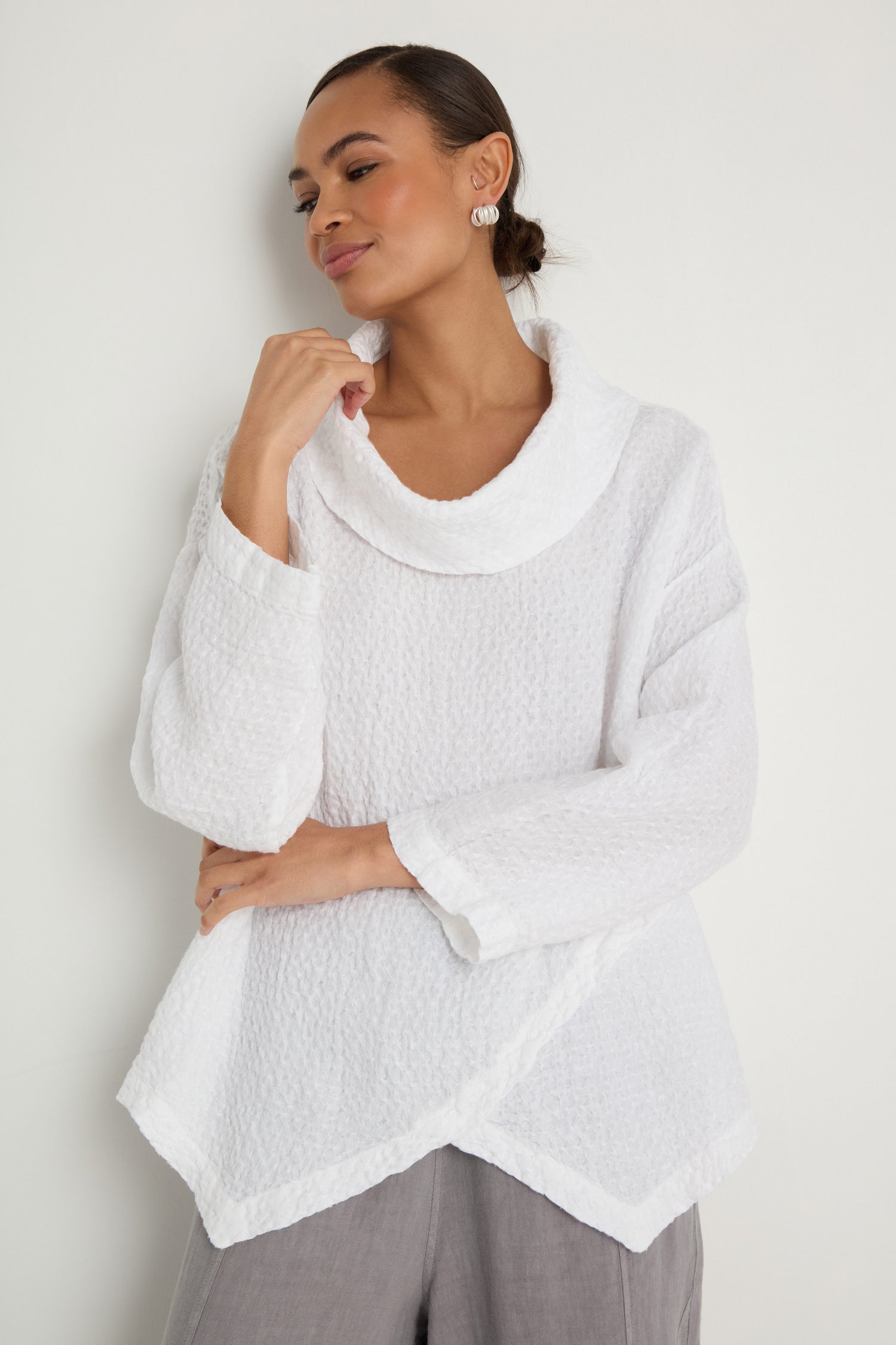 A person stands against a white wall wearing the Waffle Linen Boxy Cowl Neck Top, a textured white sweater with long sleeves, paired with light gray pants.