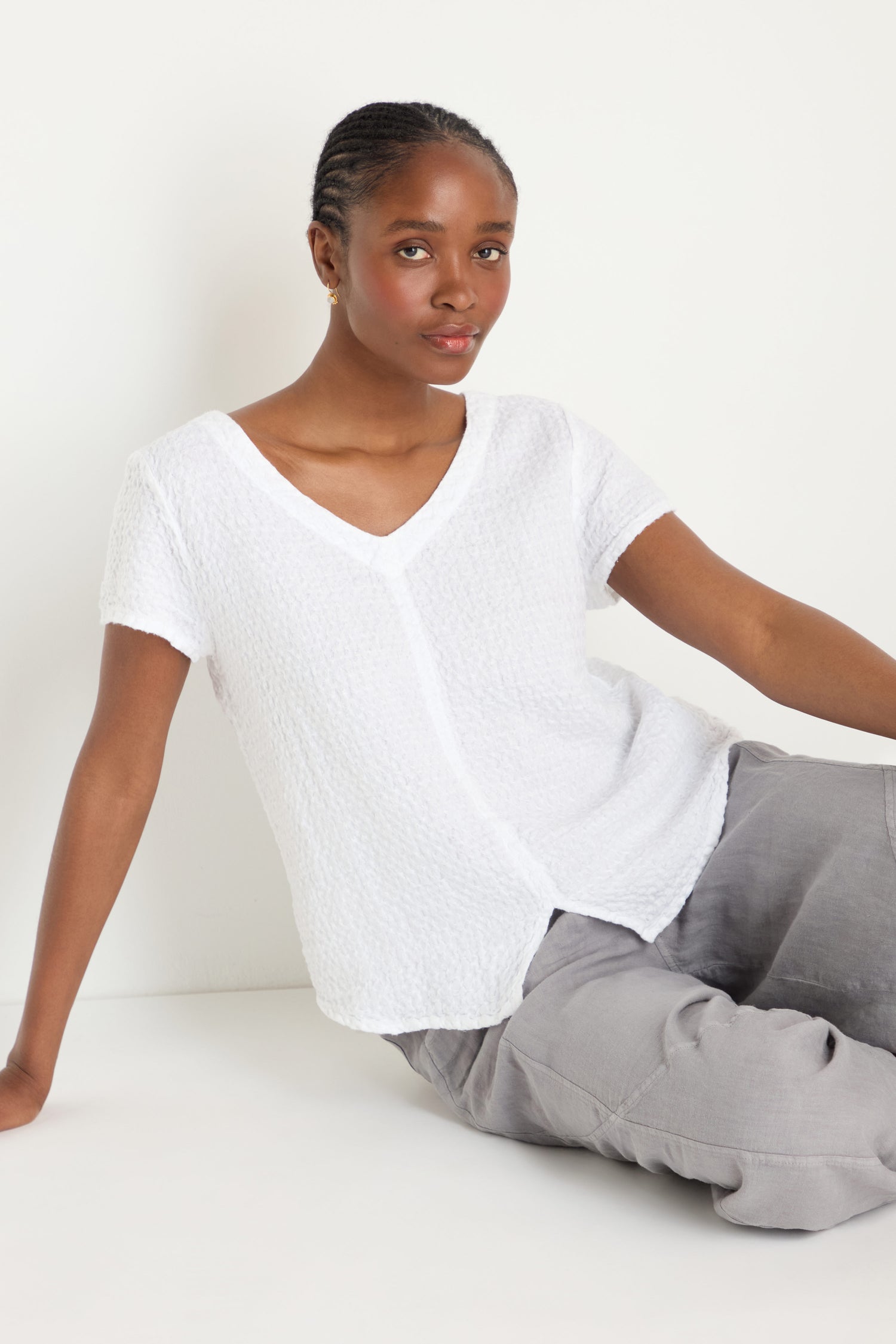 A person sits on the floor against a white wall, wearing a textured white short-sleeve Waffle Linen V Neck Top and light gray pants.