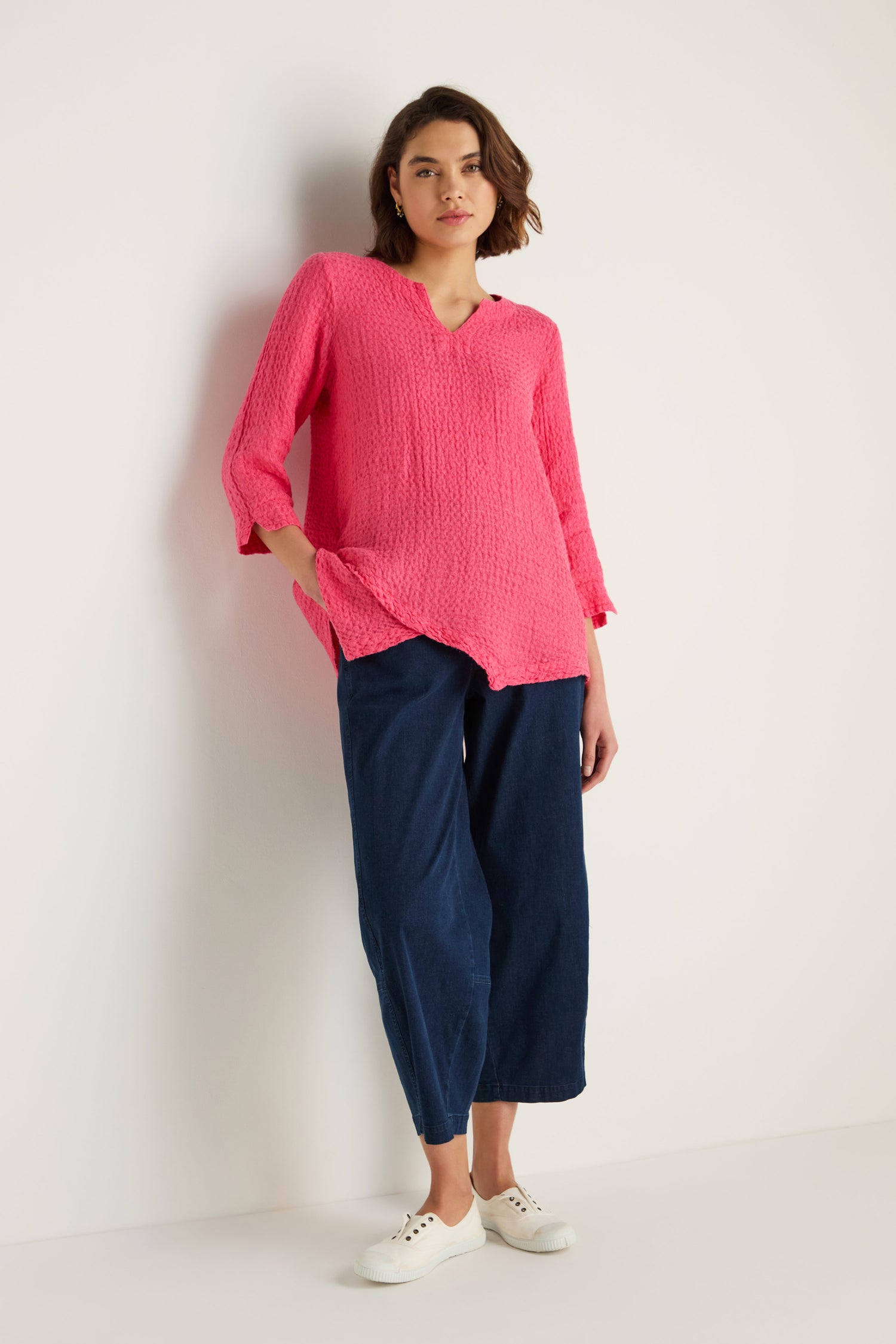 A person stands against a white wall wearing the bright pink Waffle Linen Asymmetric Tunic, navy wide-leg pants, and white lace-up shoes.