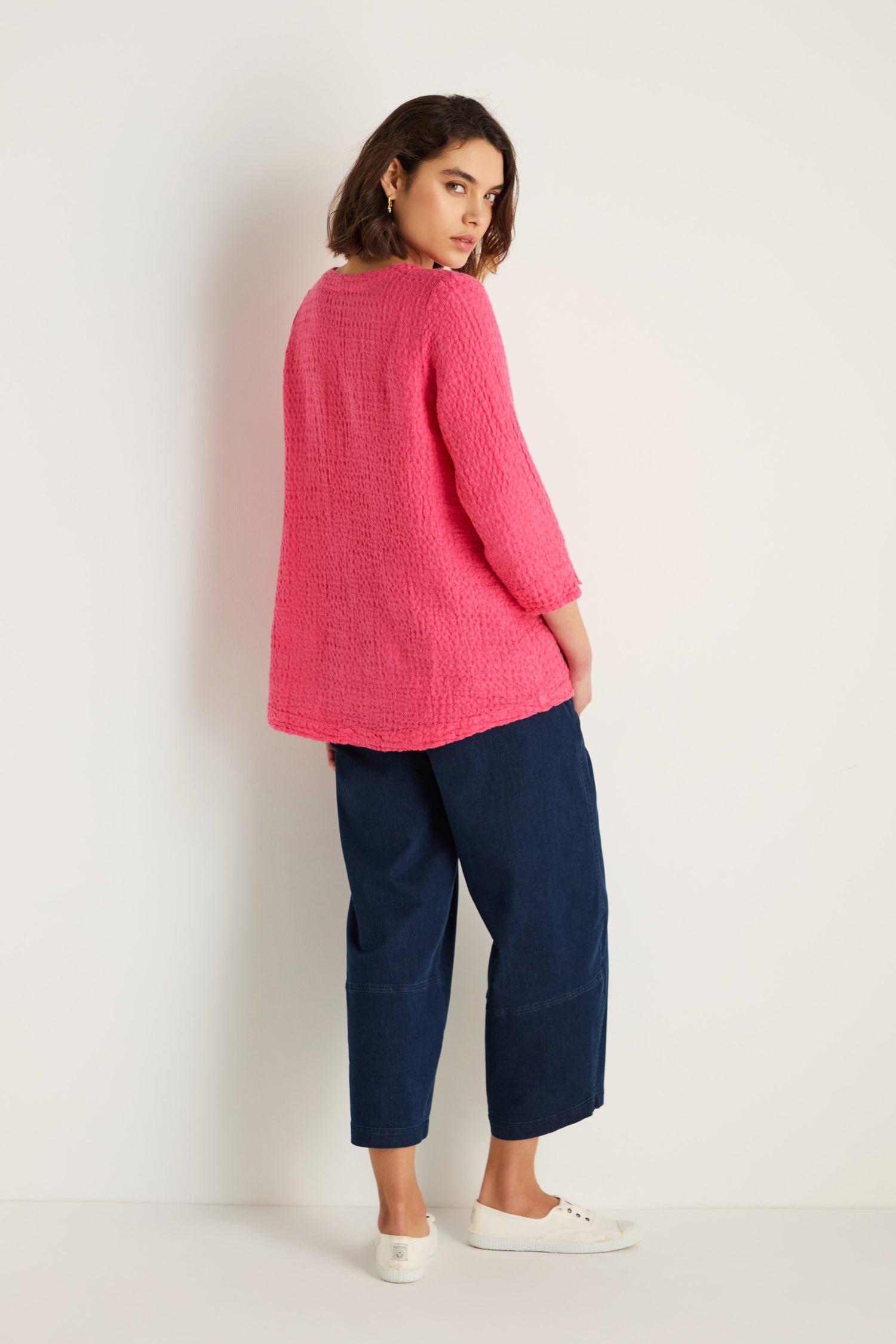 A woman faces away, glancing over her shoulder, wearing the Waffle Linen Asymmetric Tunic in pink, paired with dark blue wide-leg pants and white slip-on shoes against a plain white background.