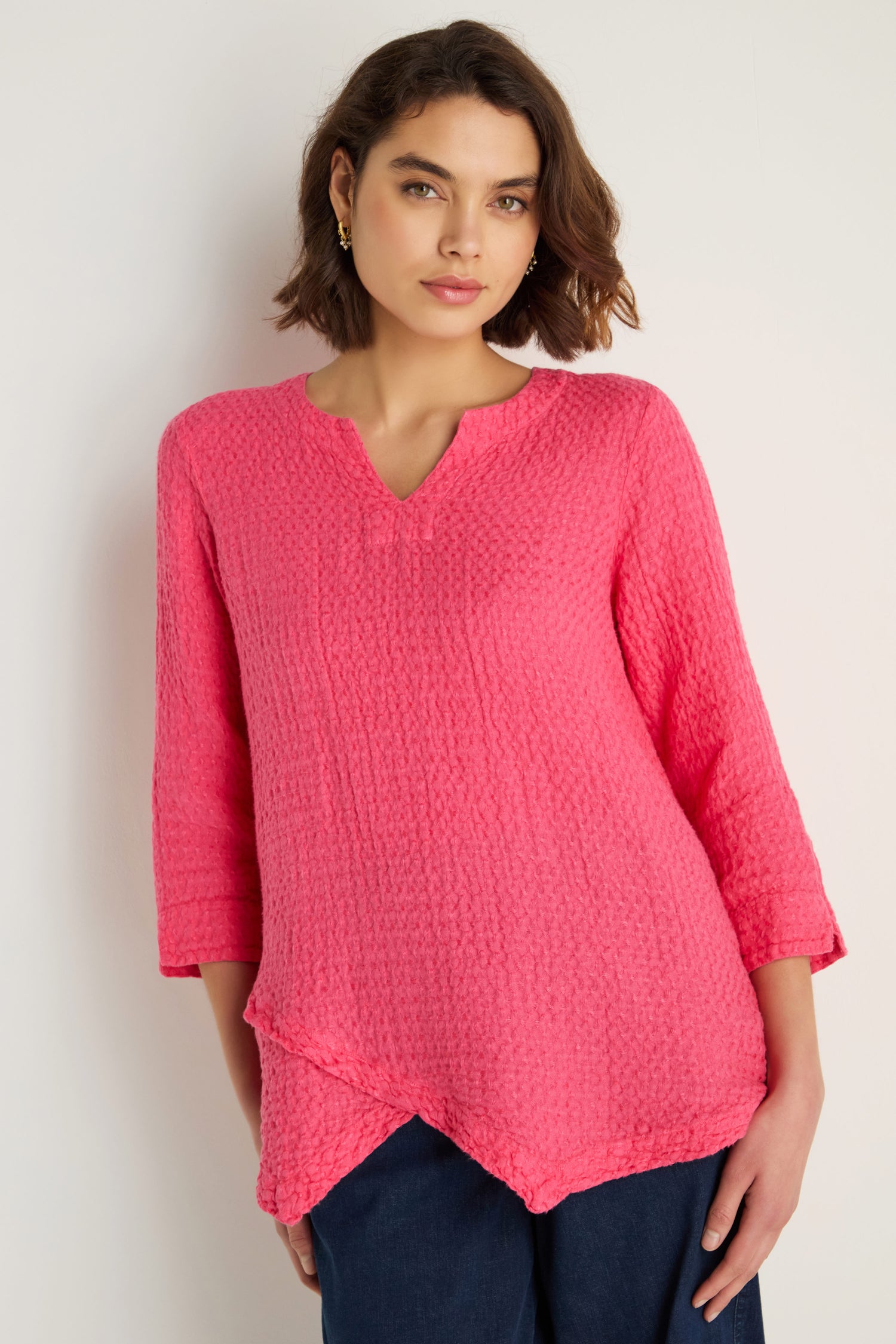 A woman models the Waffle Linen Asymmetric Tunic in bright pink, featuring a notched neckline and long sleeves, styled with dark pants against a plain light background.
