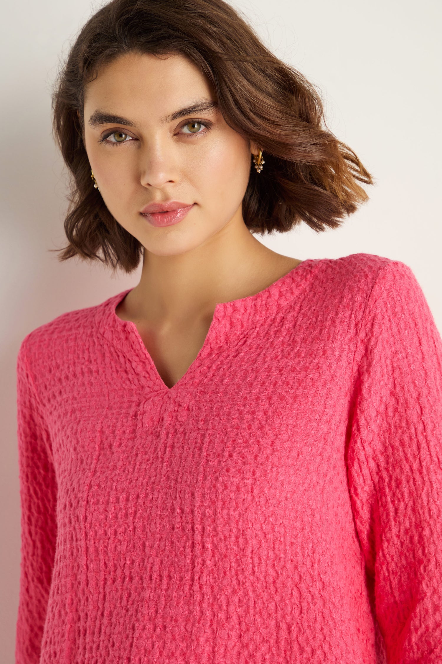 A woman with short brown hair models the Waffle Linen Asymmetric Tunic in bright pink, featuring a notched neckline and waffle texture, standing against a plain white background.