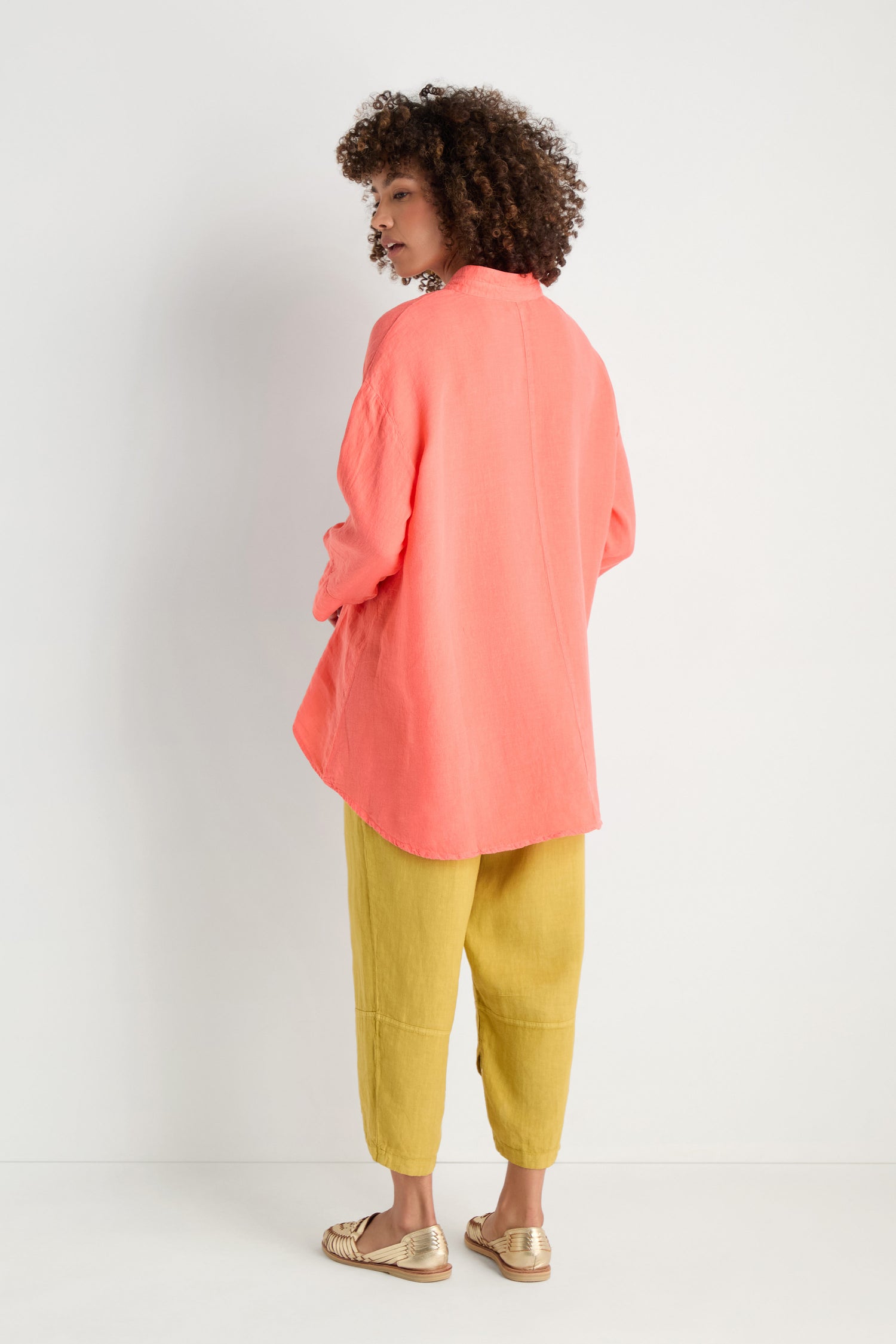 A person stands against a white wall, facing away, wearing the Organza Linen Flared Shirt in coral pink with a Nehru collar, paired with yellow pants and beige sandals.