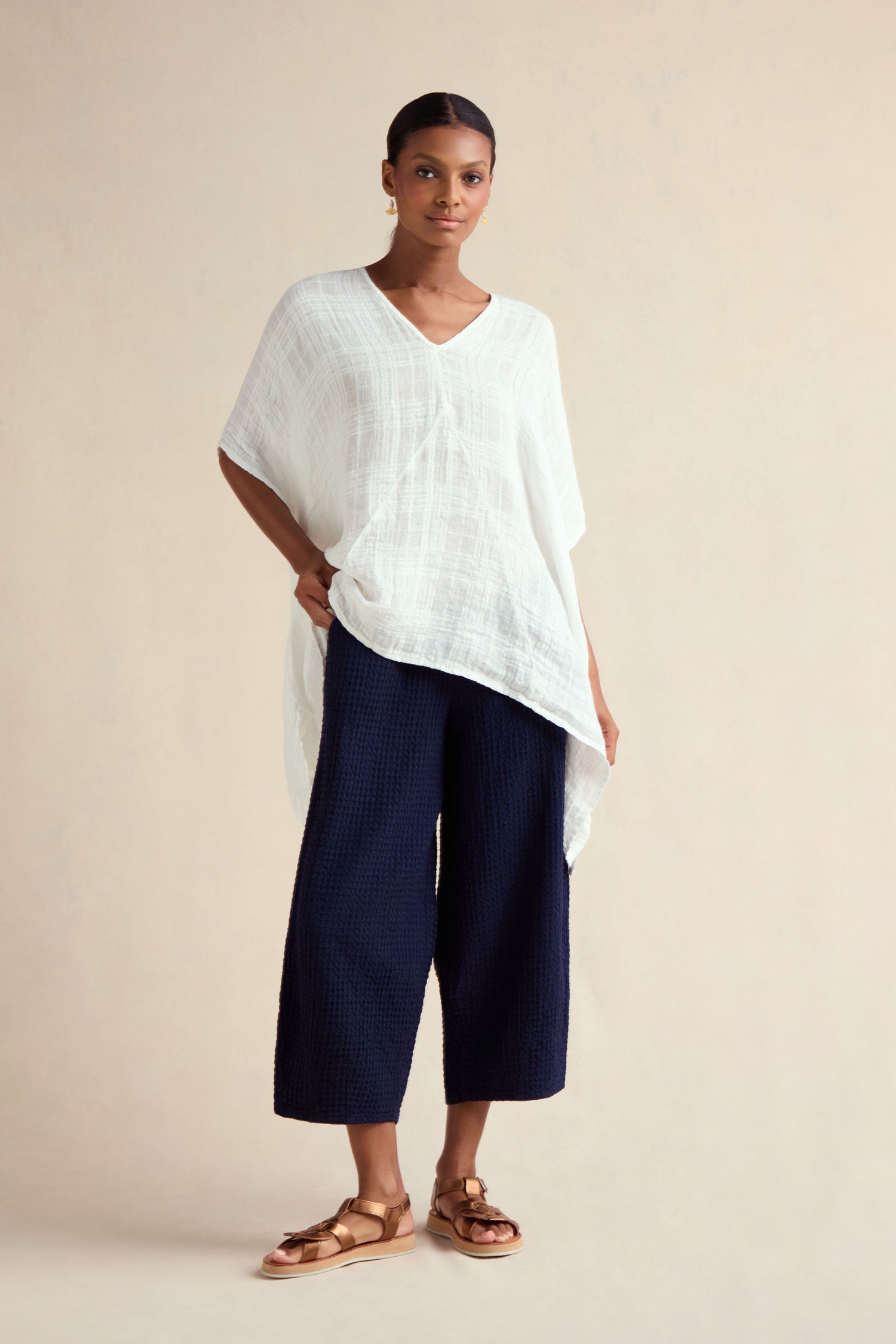A woman stands against a neutral background wearing the Gauzy Linen Boxy Tunic in white, paired with navy wide-leg textured pants and gold sandals.