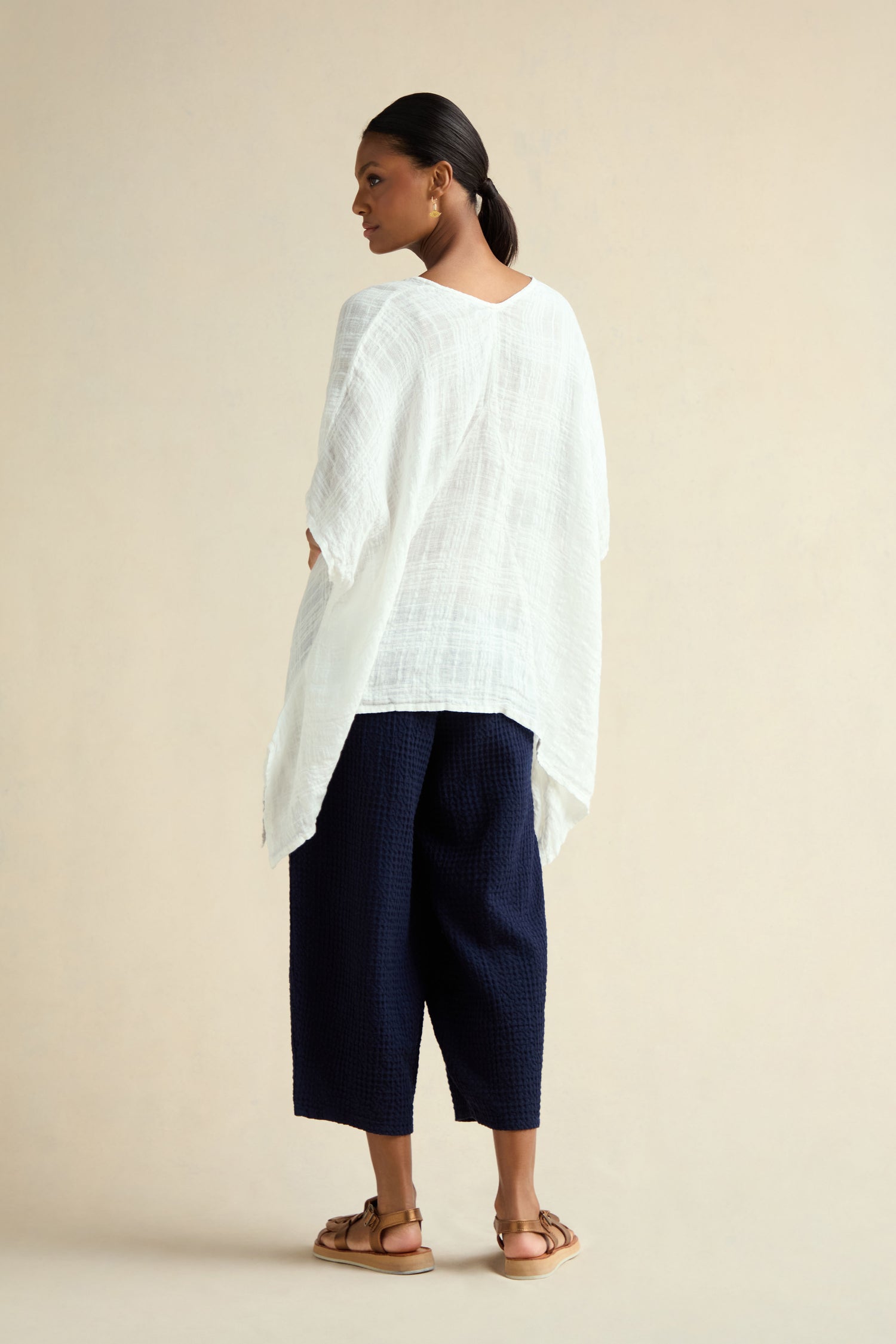 A woman stands facing away in a Gauzy Linen Boxy Tunic, navy textured wide-leg pants, tan sandals, and gold earrings against a plain beige background.