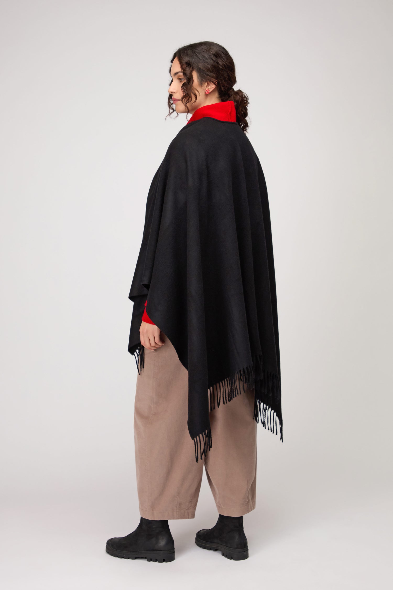 A person stands sideways against a plain background, wearing a red shirt, beige pants, black boots, and the Wool Cashmere Black Shawl with fringe.
