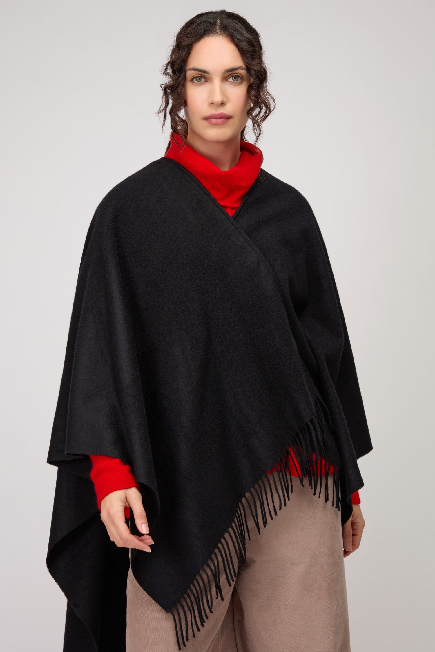 A woman stands against a plain light background, draped in the Wool Cashmere Black Shawl over a red turtleneck sweater and beige pants.