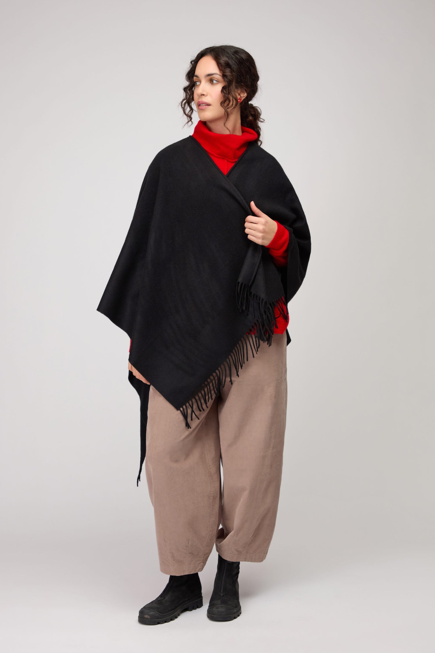 A woman stands against a plain light background wearing the Wool Cashmere Black Shawl with fringe over a red turtleneck, loose beige pants, and black boots.