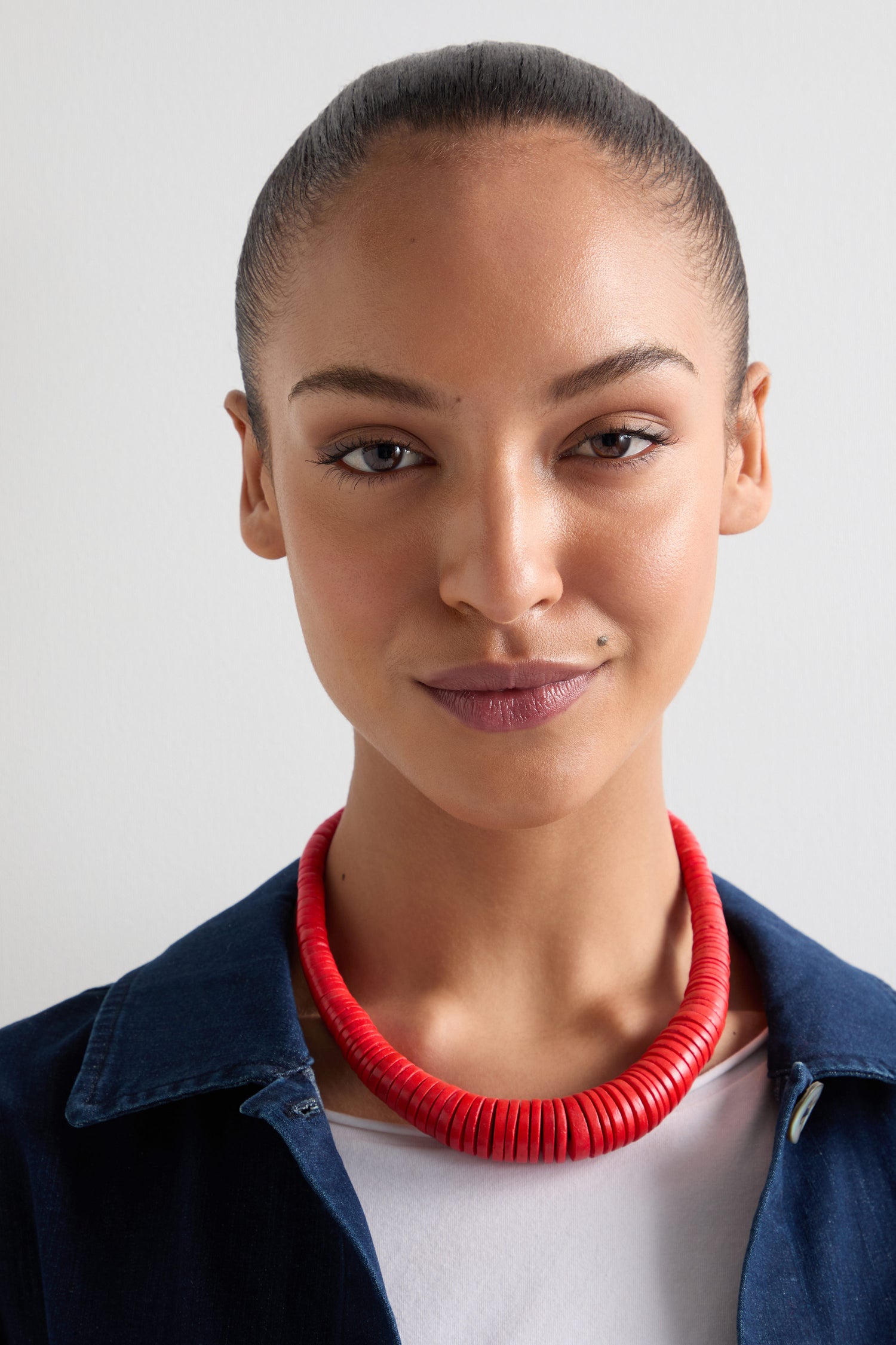 A person with tied-back hair, wearing a blue jacket over a white top and the Graduated Coco Disc Necklace—an ethically sourced, red beaded piece showcasing exquisite Filipino craftsmanship—stands against a plain background.