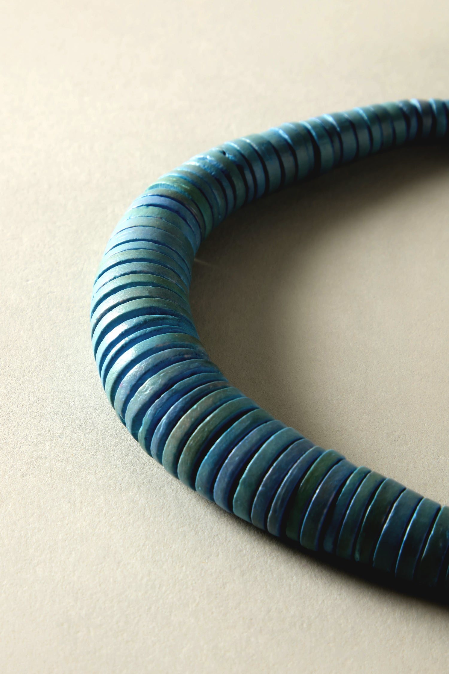 A close-up of the Graduated Coco Disc Necklace, ethically sourced with blue beads, exhibits exquisite craftsmanship in a semi-circle on a neutral background.
