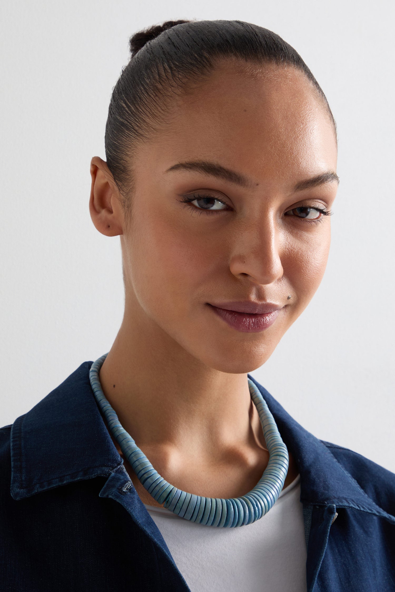A person with their hair tied back wears a blue Graduated Coco Disc Necklace, ethically sourced, and a dark collared shirt while looking at the camera against a plain background.