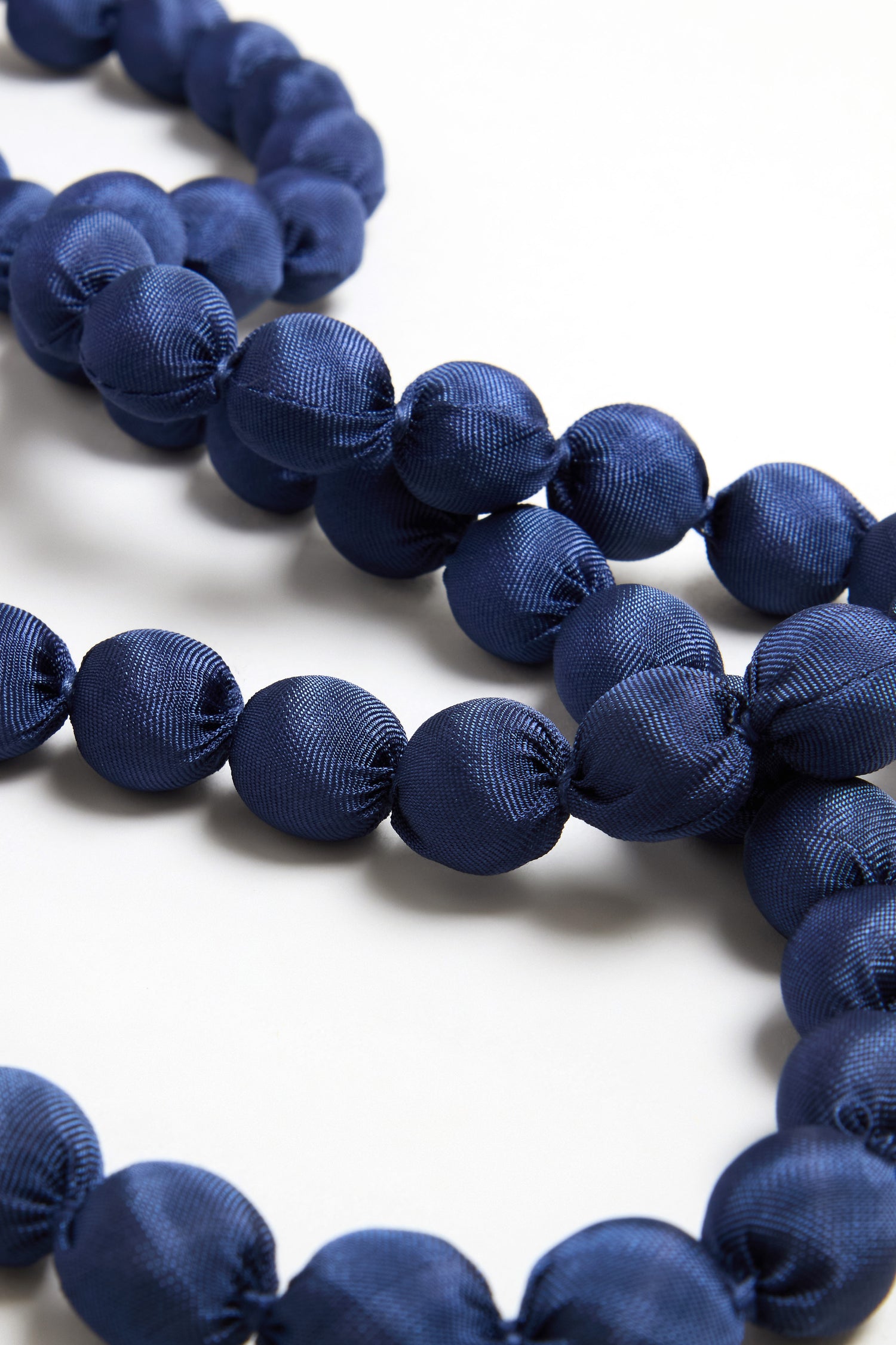A close-up of the Handmade Long Silk Spheres Necklace showcases large, round, navy blue silk-wrapped beads arranged in strands on a white surface.