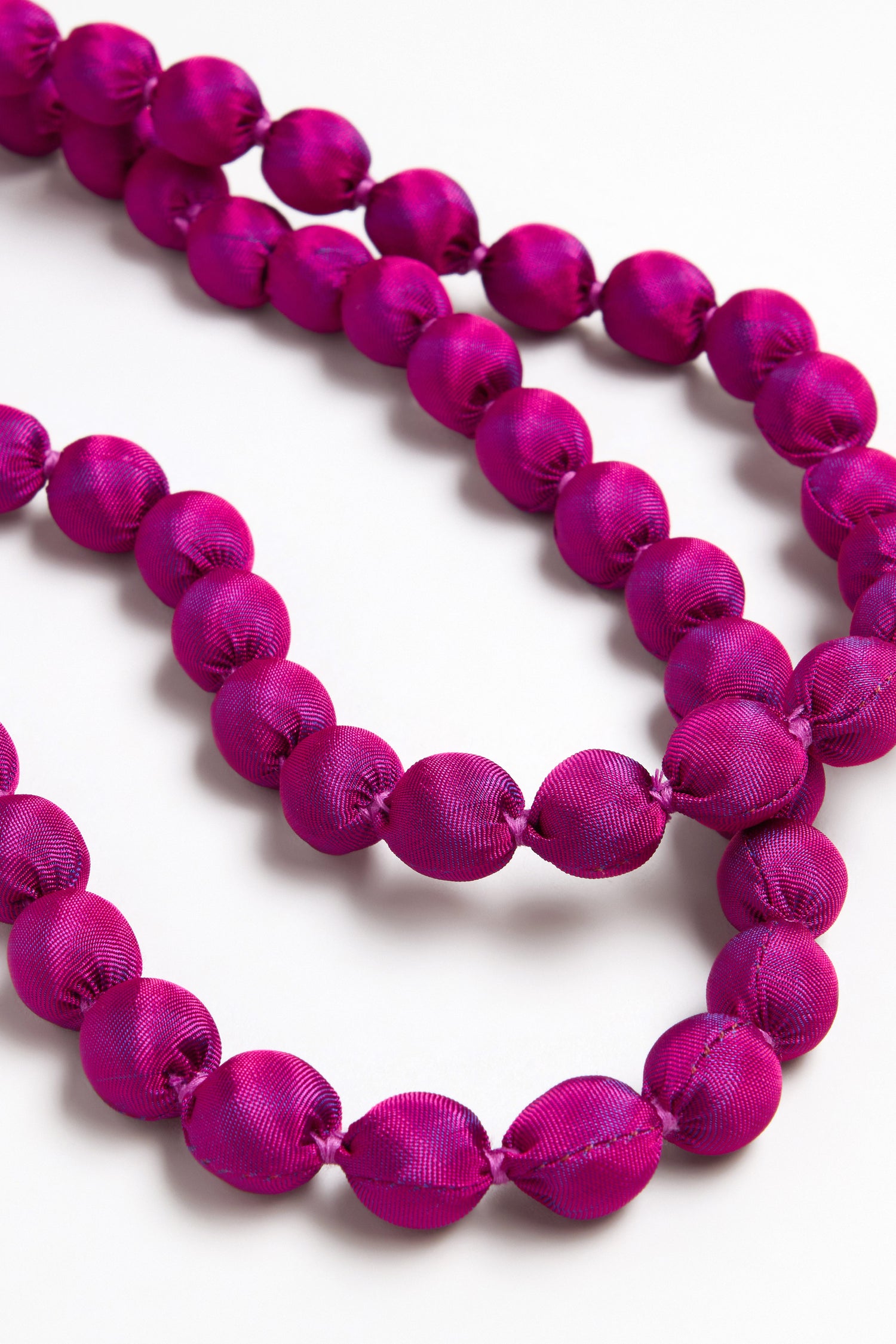 Close-up of the Handmade Long Silk Spheres Fuchsia Necklace, featuring round beads covered in fuchsia satin fabric and arranged in loose loops on a white background.