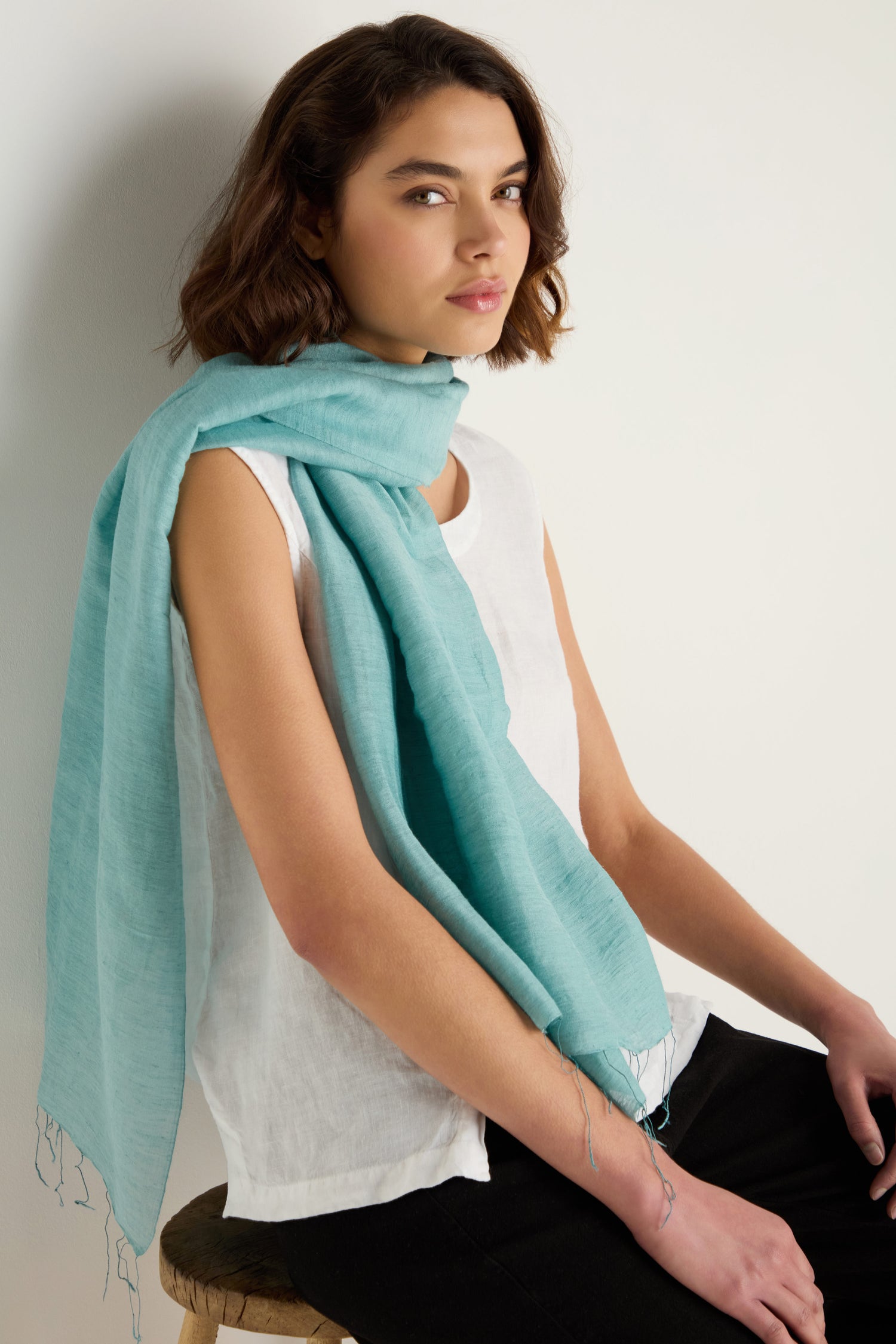 A woman with short brown hair wears a sleeveless white top, black pants, and the Soft Linen Fringe Scarf while sitting on a wooden stool against a plain white background.