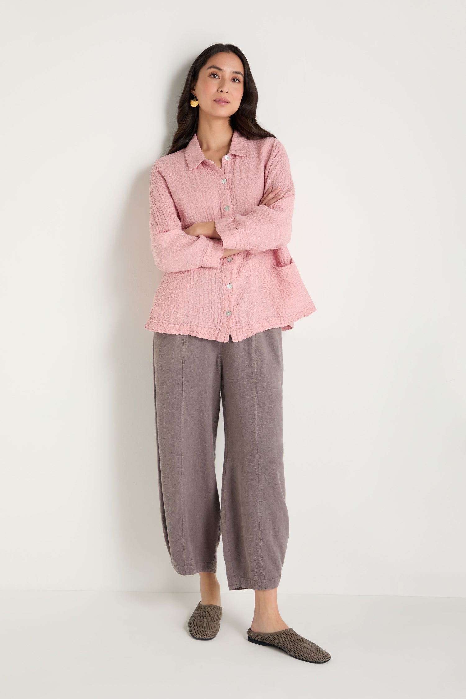A woman stands against a white wall, wearing a light pink textured button-up shirt, loose gray pants, and Mesh Leather Slip on Shoes handmade in Europe. Her arms are crossed as she faces forward.