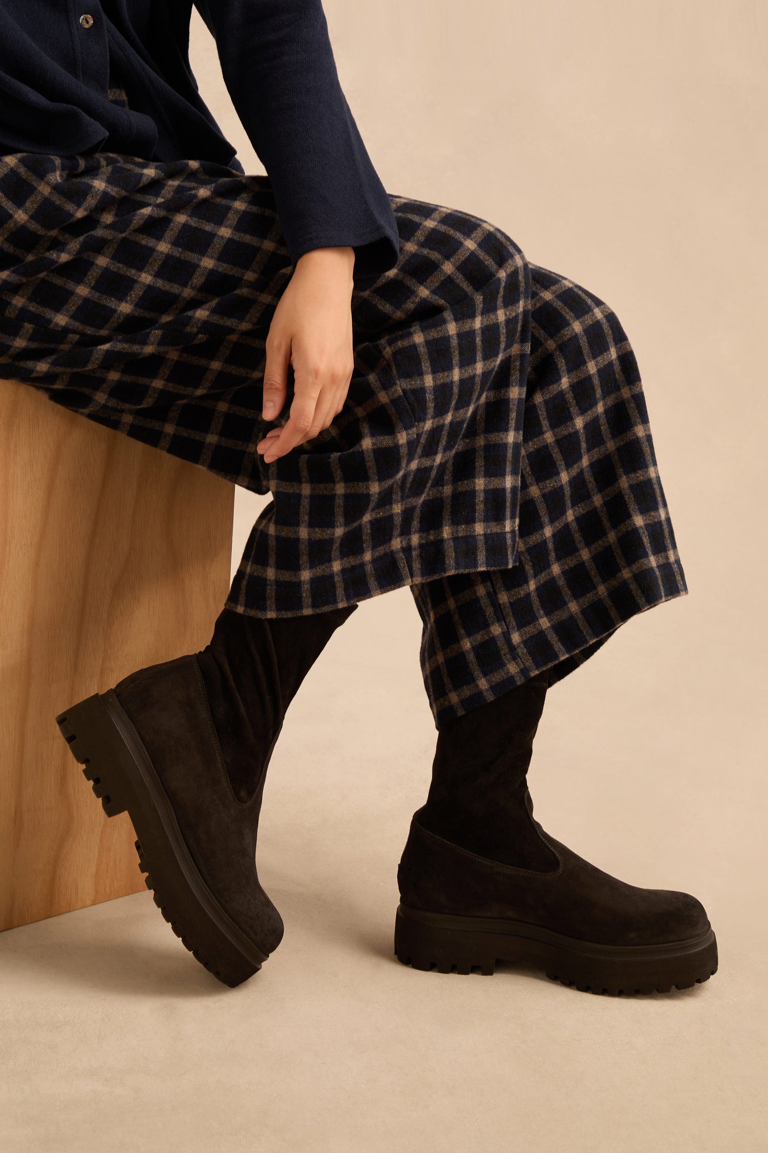 Person sitting on a wooden block, wearing a navy top, plaid pants, and Short Stretch Suede Boots with chunky soles against a beige background.