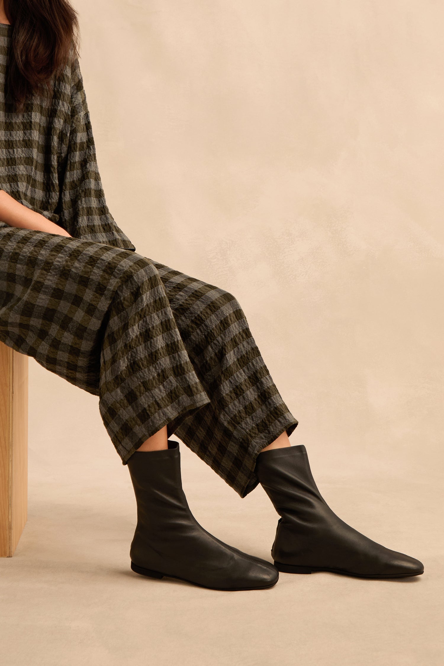 A person sits on a wooden bench wearing a black and gray checkered outfit and sleek Slim Napa Leather Stretch Boots, against a beige background.