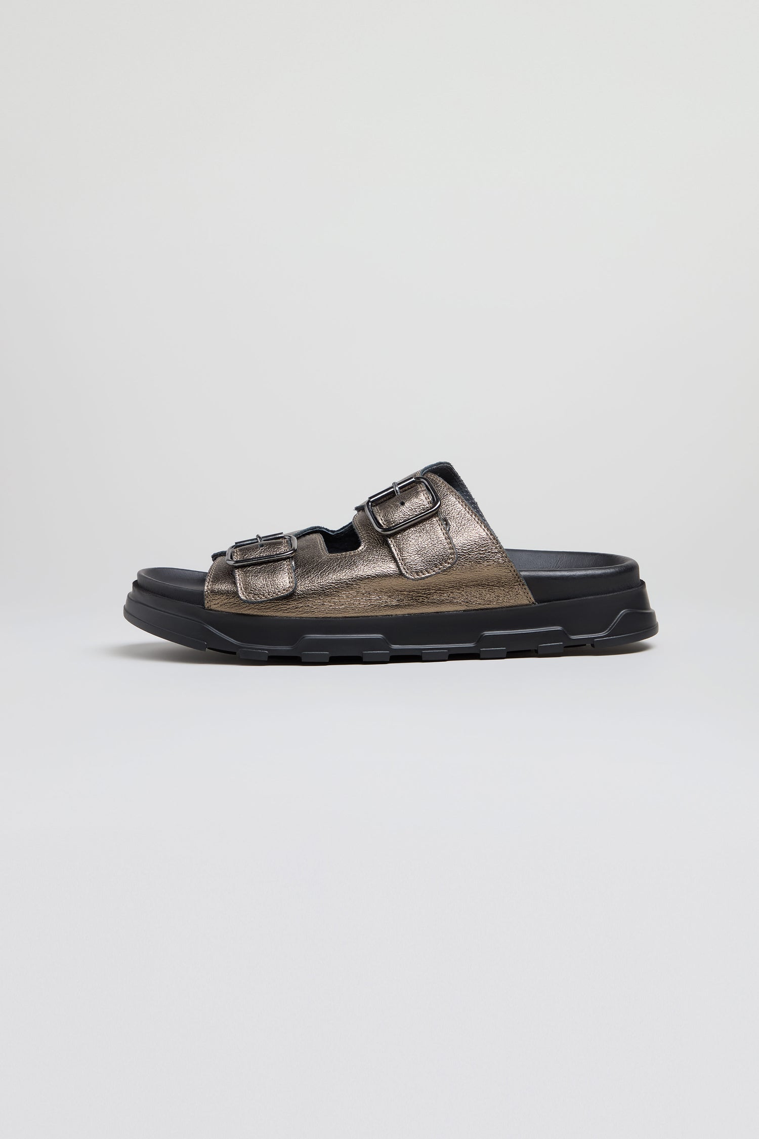 The Double Buckle Leather Sliders are handcrafted in Italy from metallic calf leather, featuring two adjustable buckle straps and a thick black sole, shown in profile against a plain background.