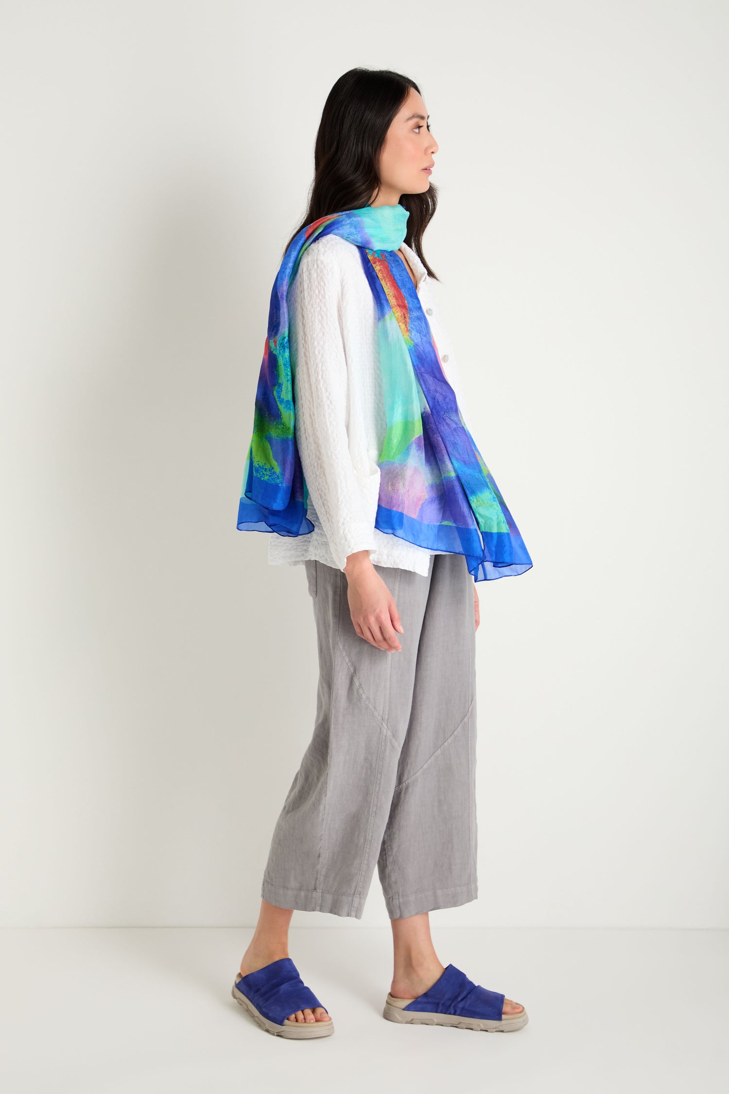 Woman standing sideways against a white wall, wearing a white textured long-sleeve top, gray cropped pants, Crushed Suede Sliders, and a colorful blue scarf draped over her shoulders.