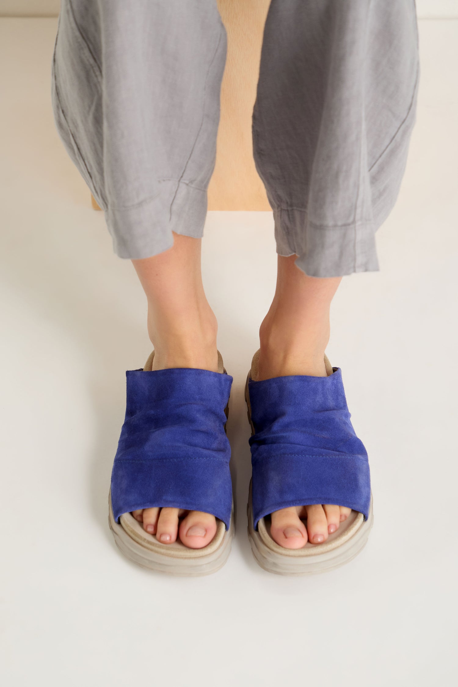 A person stands on a light surface wearing loose gray pants and blue Crushed Suede Sliders made in Italy.