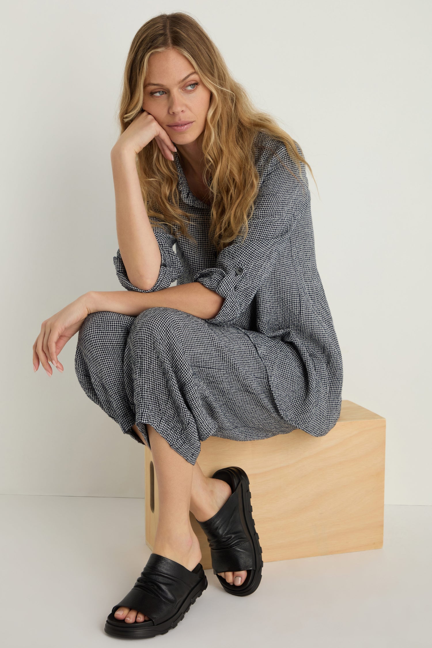 A woman with long wavy hair sits on a wooden box, wearing a gray checked outfit and Crushed Metallic Leather Sliders handcrafted in Italy, resting her head on her hand as she looks to the side.