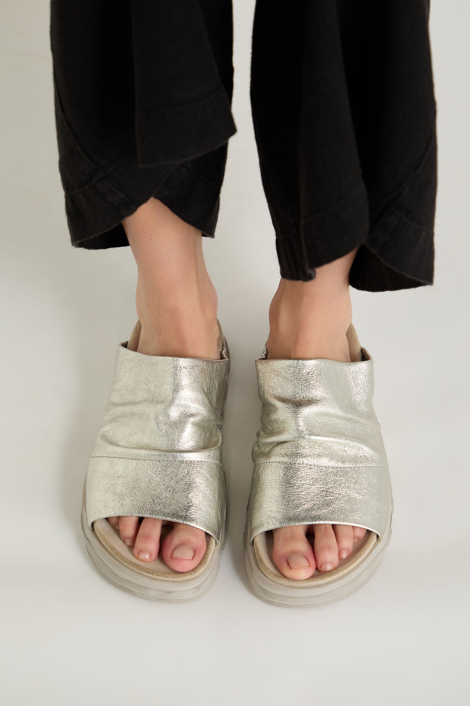 A person wearing wide-legged black pants and Crushed Metallic Leather Sliders—handcrafted women's luxury sandals—stands on a light-colored surface.
