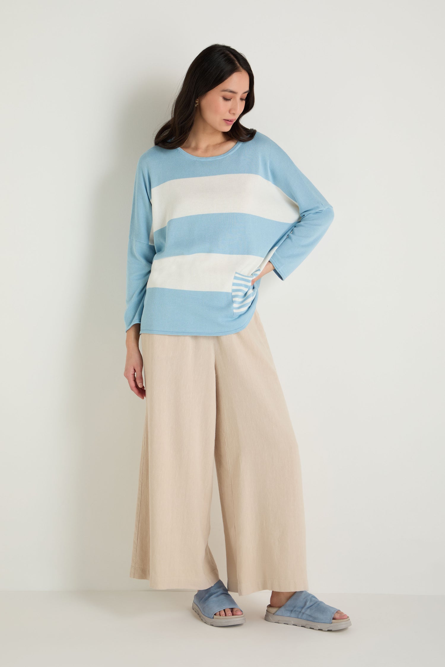 A woman stands against a plain background wearing a light blue and white striped sweater, wide-leg beige pants, and Crushed Suede Sliders, with one hand in her pocket.