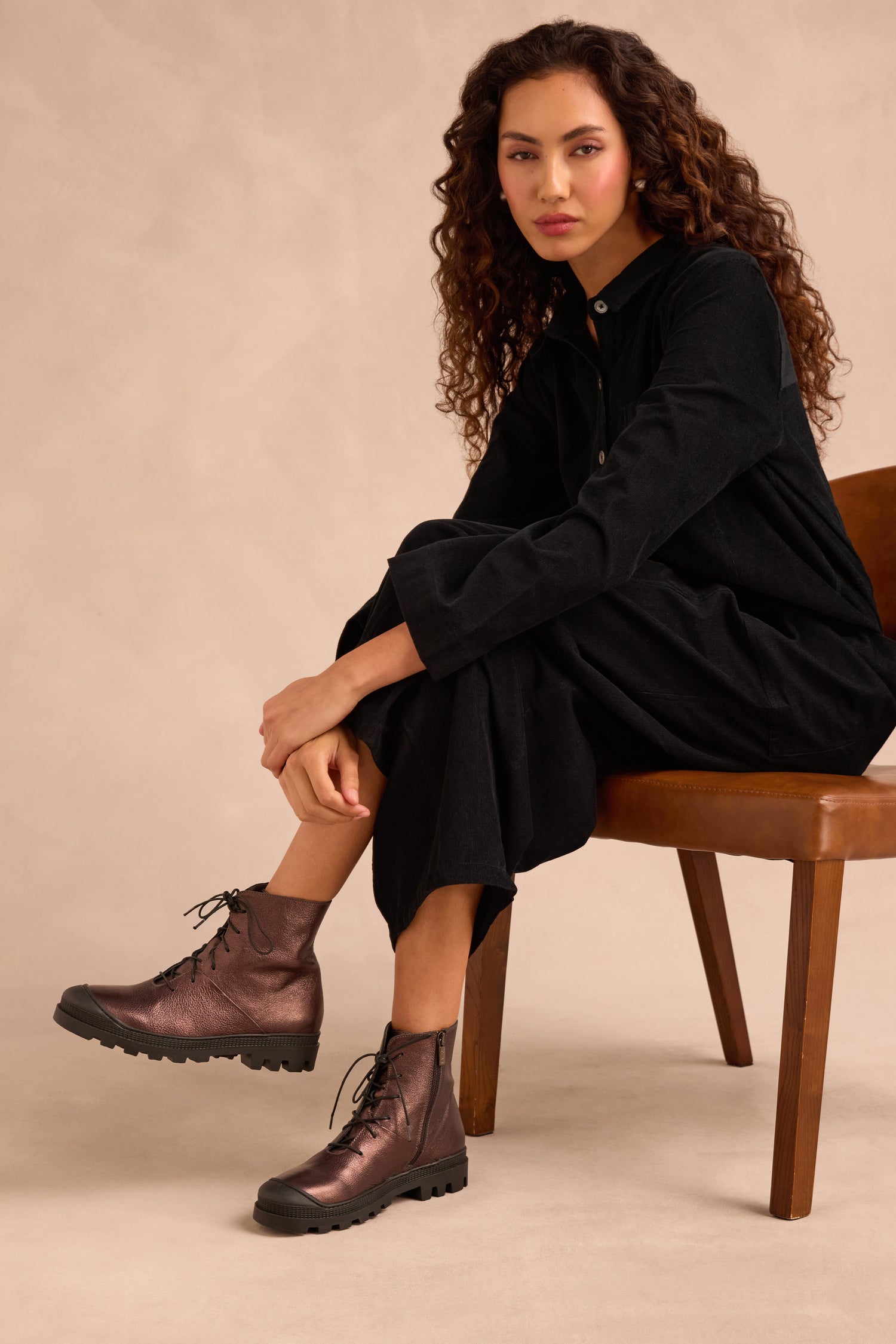 A woman sits on a chair wearing a long black dress and Metallic Chocolate Lace Up Leather Boots, looking at the camera against a neutral background.