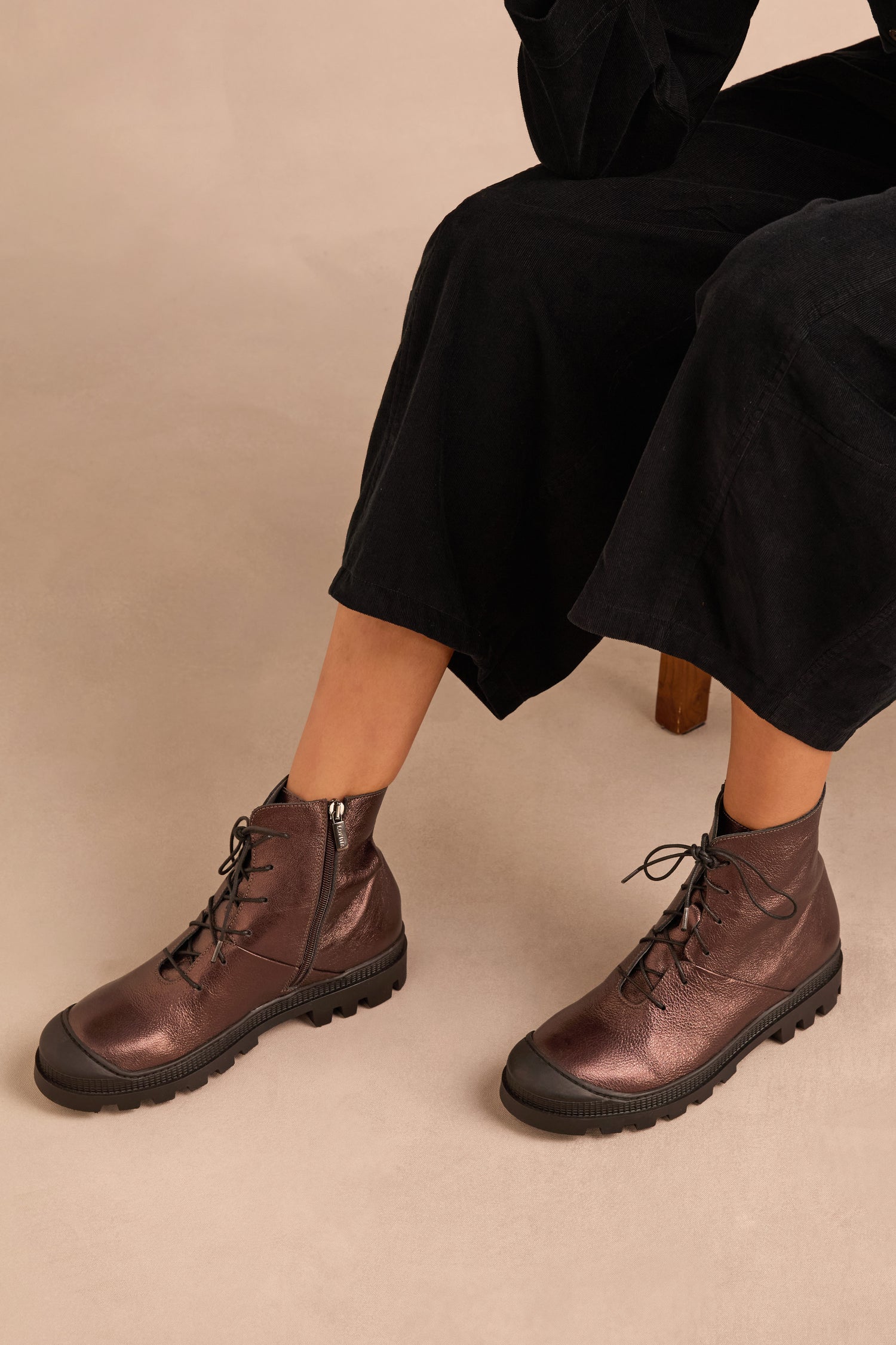 A person wearing Metallic Chocolate Lace Up Leather Boots with side zippers, seated on a chair in a black outfit against a beige background.