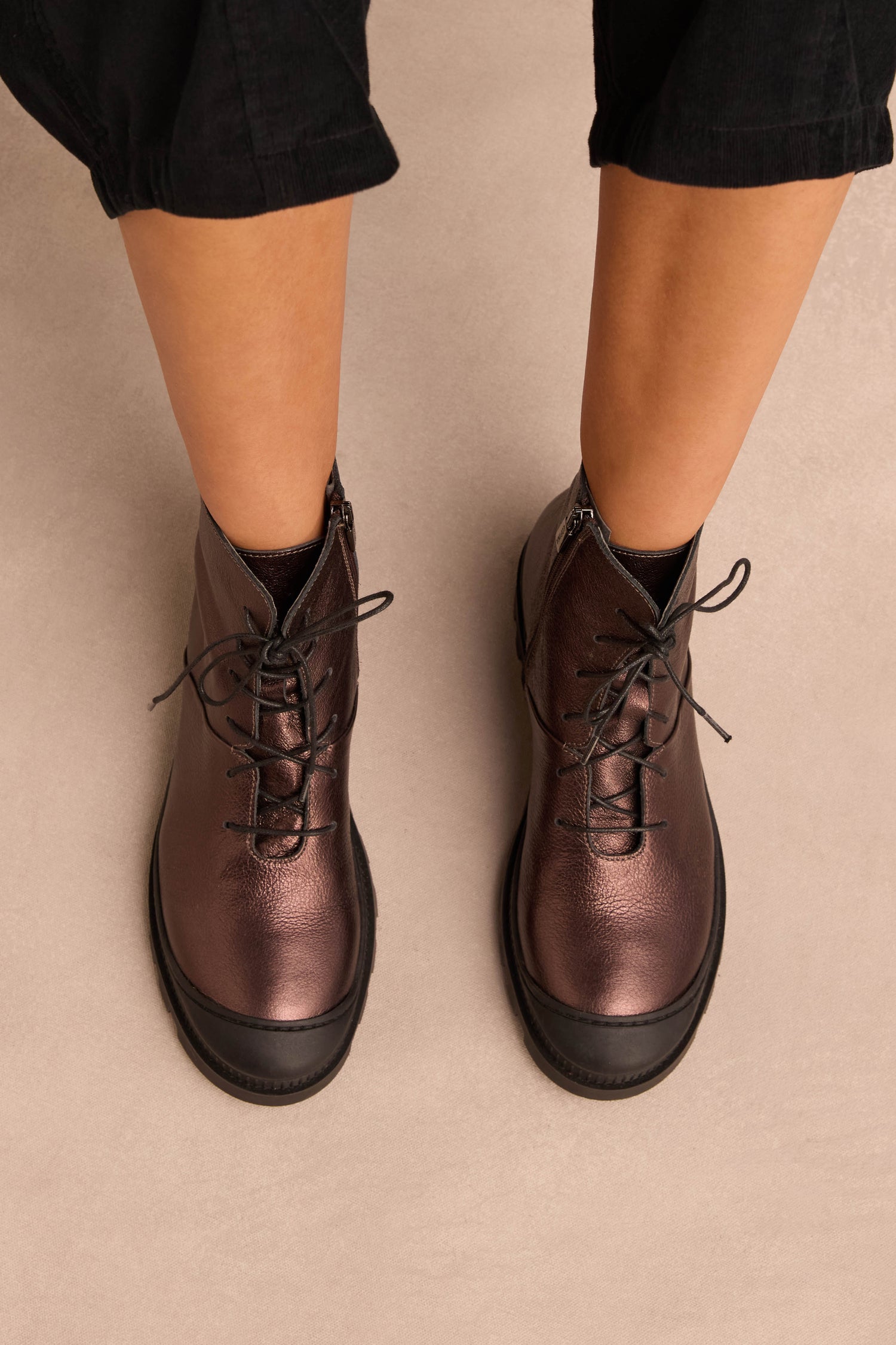Person wearing black pants and Metallic Chocolate Lace Up Leather Boots, crafted from supple calfskin, standing on a light beige surface.