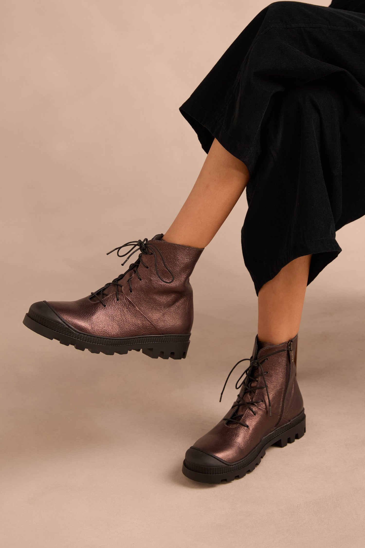 Person wearing black pants and Metallic Chocolate Lace Up Leather Boots, crafted in calfskin with thick rubber soles, seated against a neutral background.