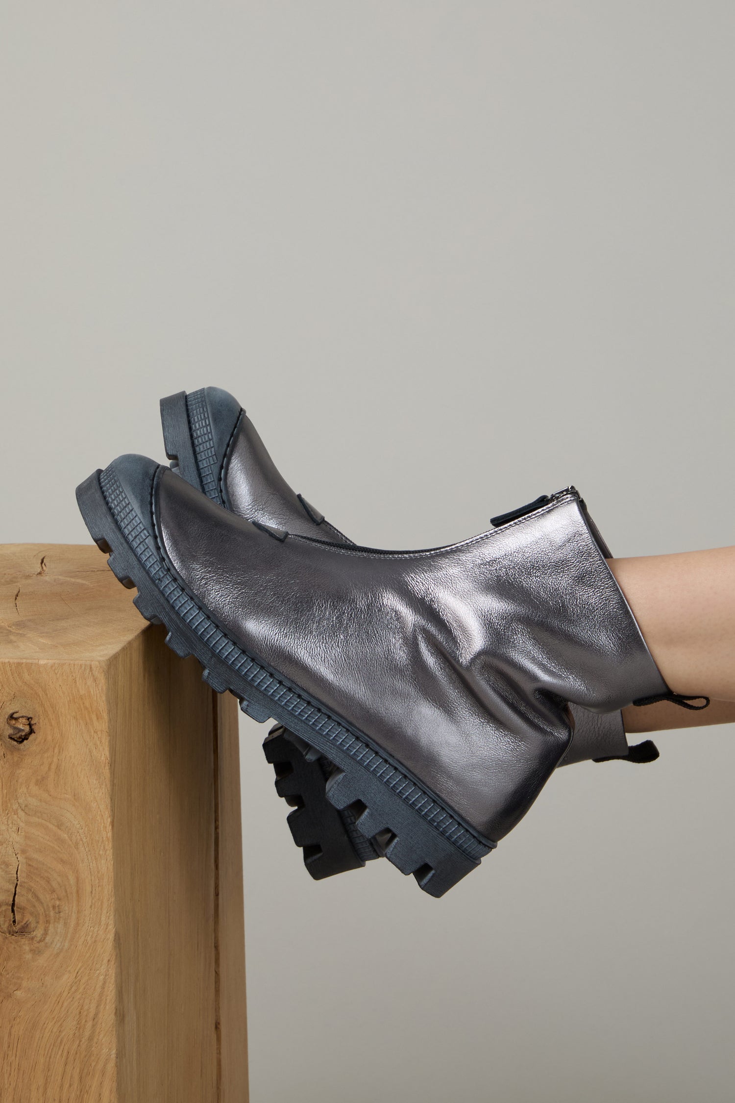 A person wearing Gunmetal Zip Front Leather Boots—shiny gunmetal ankle boots with chunky blue soles—rests their feet on a light wood block against a plain background.