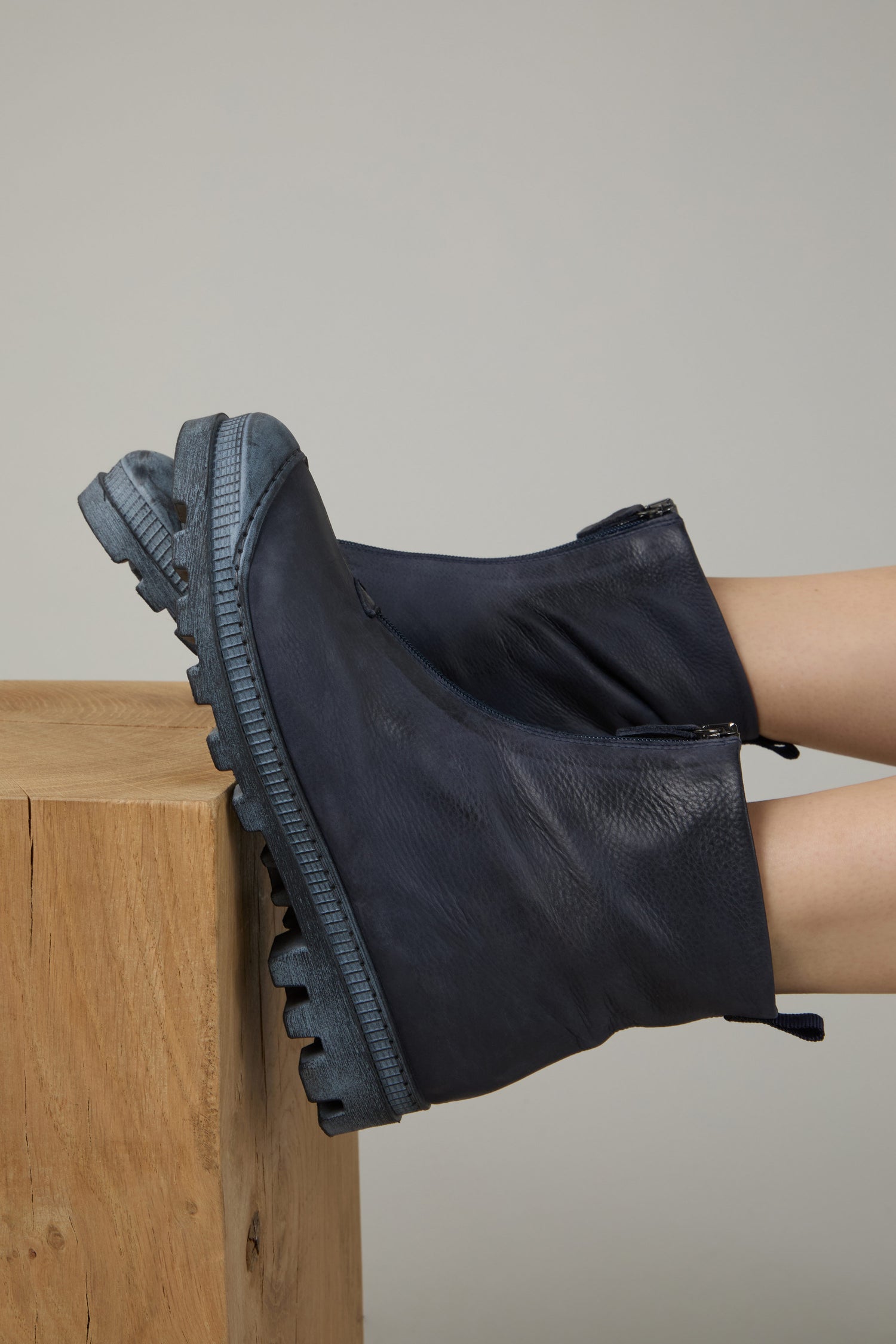 Wearing Navy Zip Front Leather Boots with thick, textured soles, a person rests their feet on a wooden block against a neutral background.