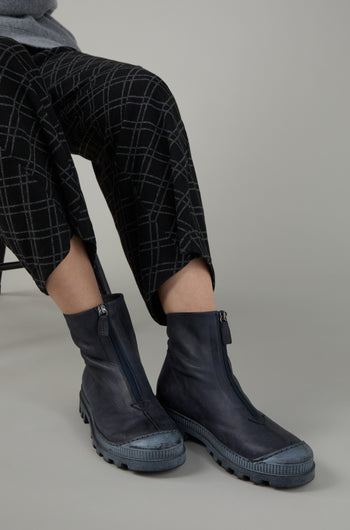 A person in black patterned pants wears Navy Zip Front Leather Boots while seated on a chair against a neutral gray background.