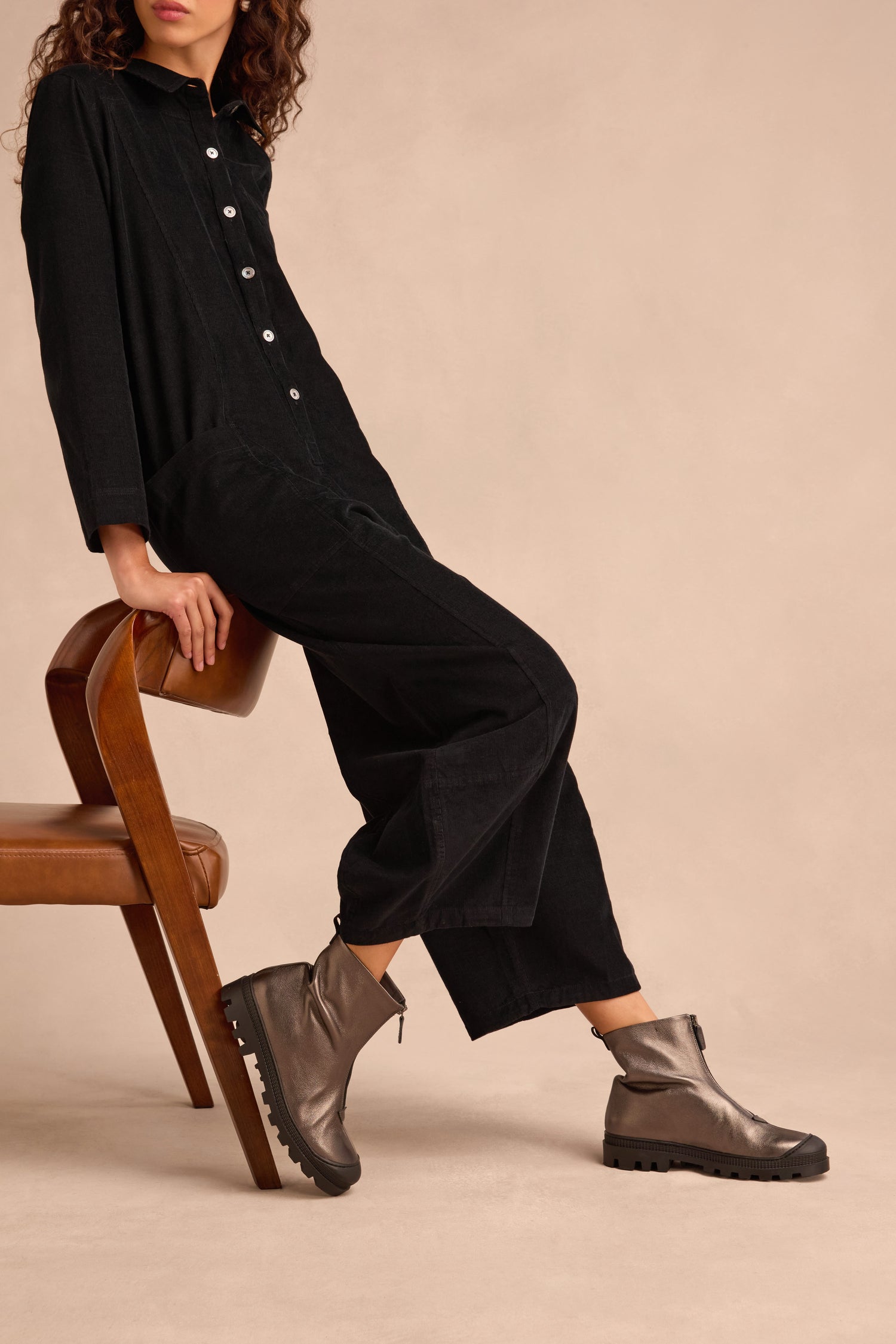 Person sits sideways on a wooden chair wearing a black long-sleeve jumpsuit and the Metallic Hematite Zip Front Leather Boots, which feature a chunky tread sole, set against a neutral background.
