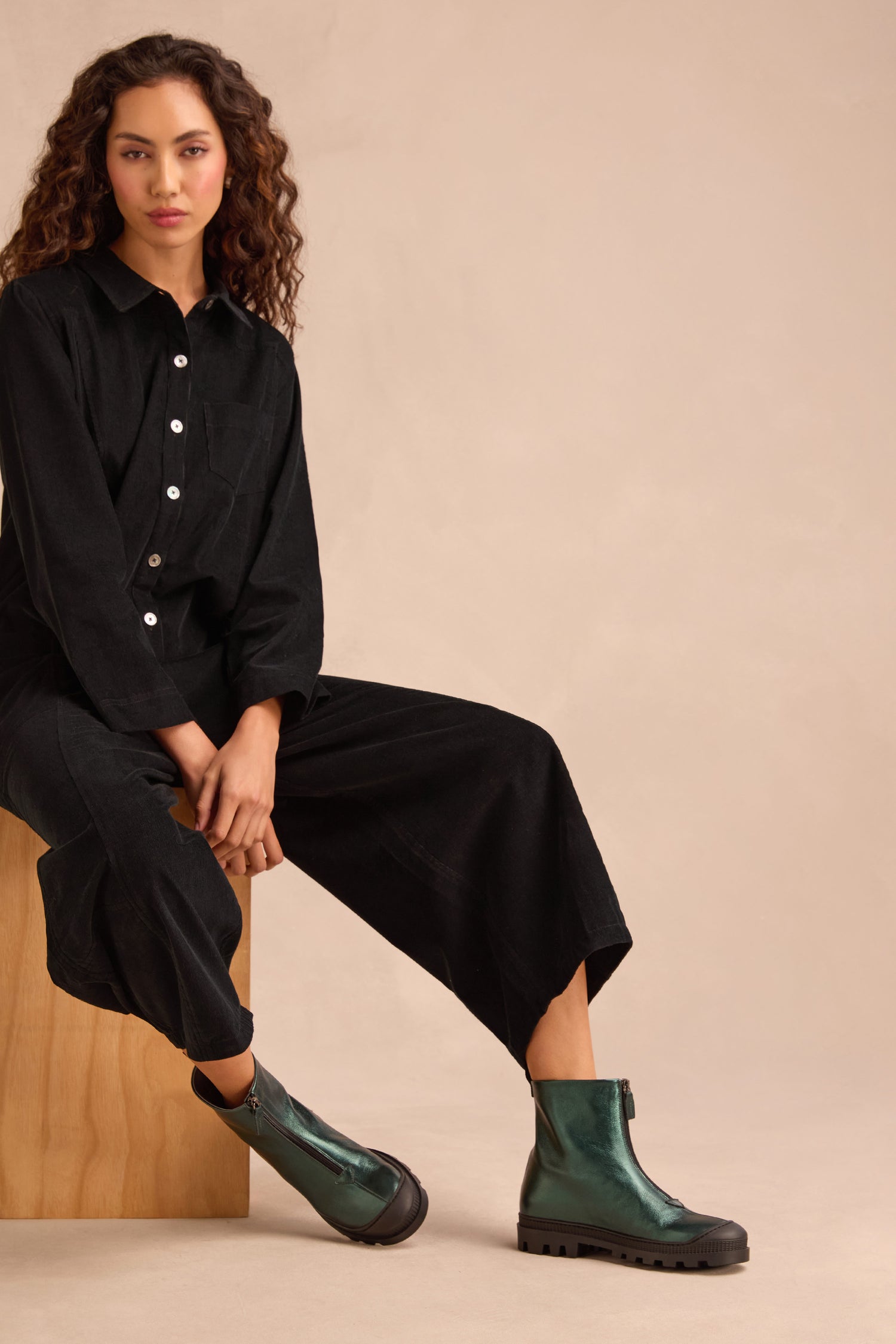 Woman sitting on a wooden block wearing a black button-up jumpsuit and the Metallic Forest Zip Front Leather Boots with a chunky tread sole, against a plain beige background.