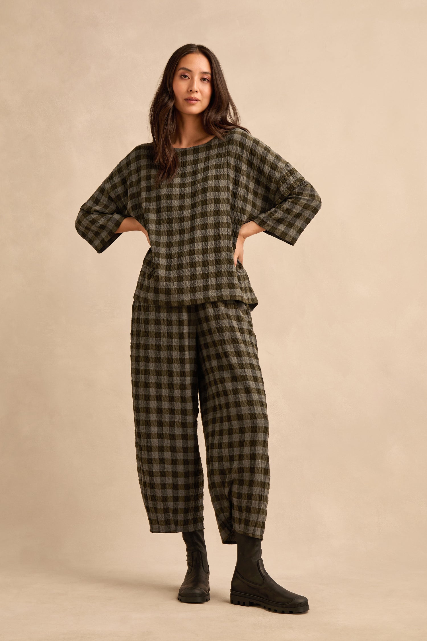 A woman stands against a plain beige background wearing a loose-fitting matching checkered top and pants, paired with Stitch Detail Napa Leather Stretch Boots.