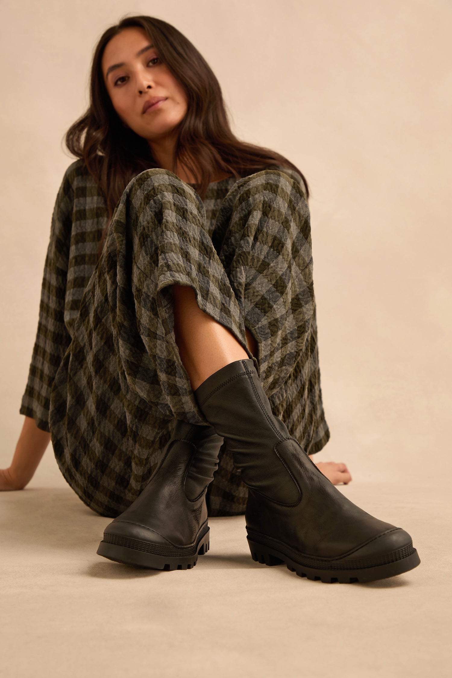 A woman in a plaid dress sits on the floor with one knee up, highlighting the Stitch Detail Napa Leather Stretch Boots—sleek, handmade in Italy—against a neutral background.