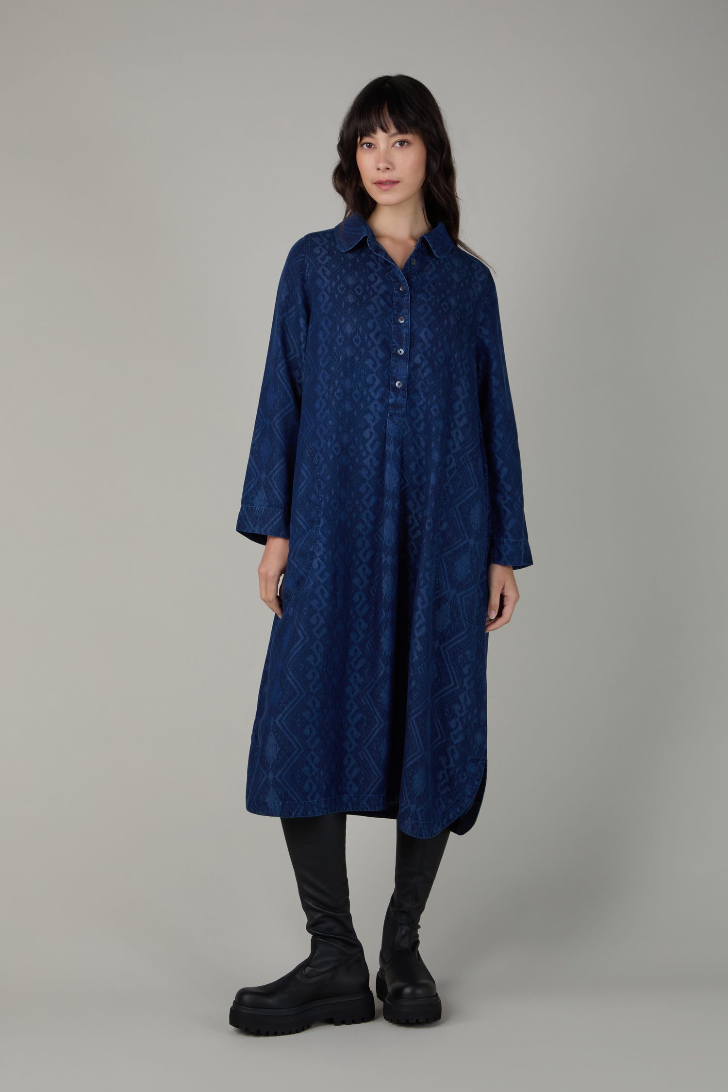 A woman stands facing forward, wearing a long-sleeved, button-up dark blue patterned dress with black leggings and Black Long Stretch Knee High Leather Boots against a plain background.