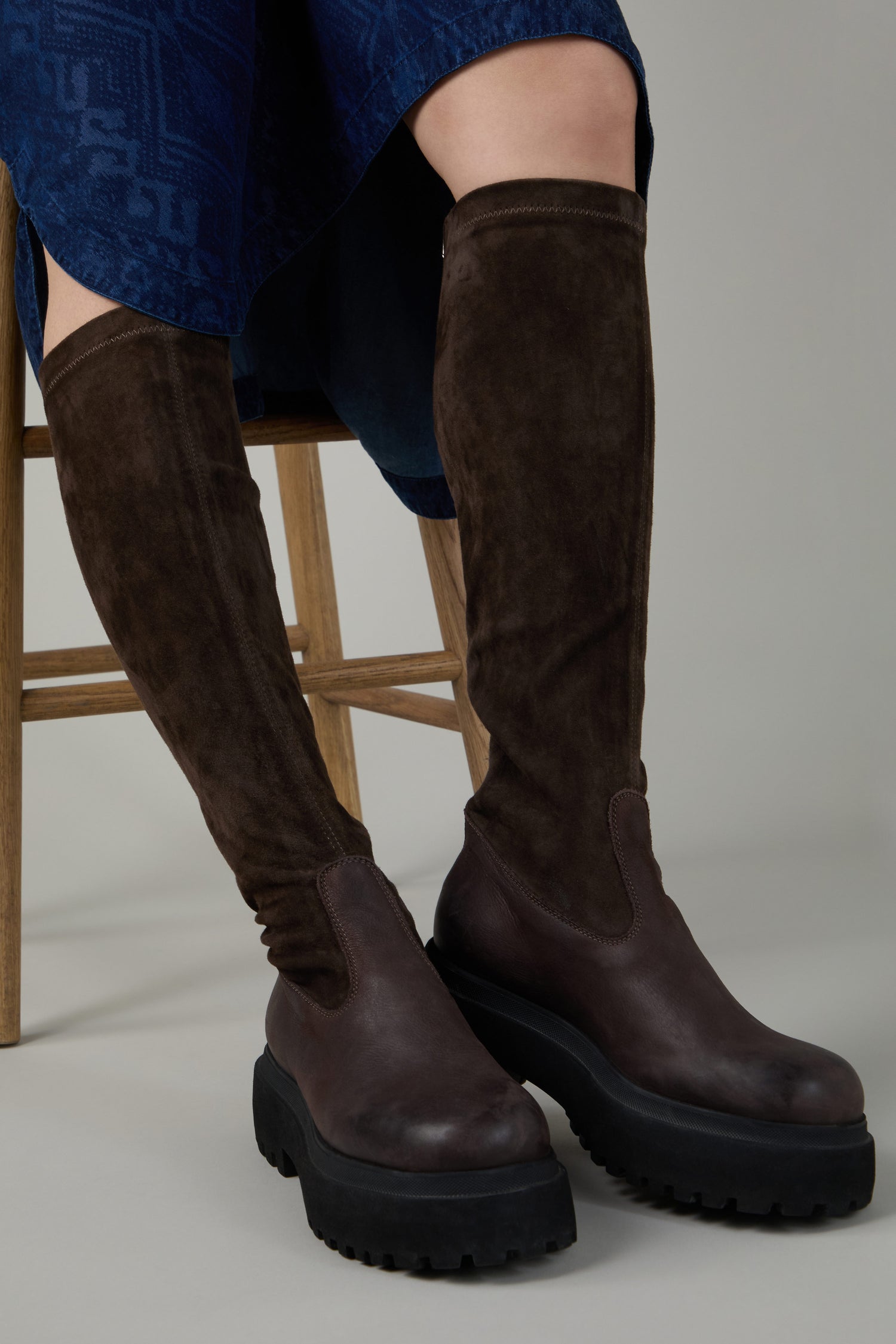 A person sits on a wooden chair, legs slightly apart, wearing Chocolate Long Stretch High Knee Suede Boots with a thick platform sole and a blue skirt visible.