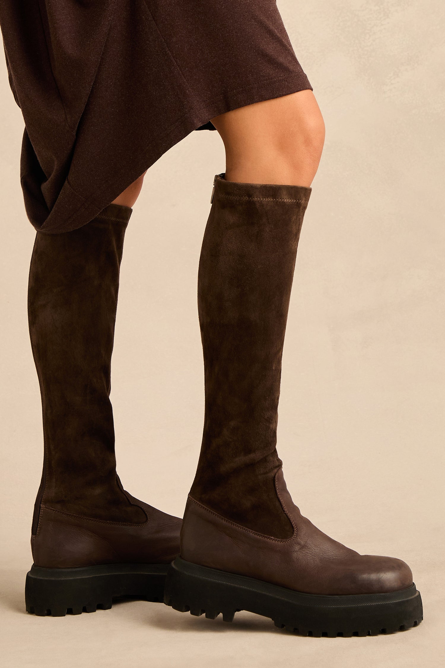 A person wearing a brown skirt and Knee High Stretch Suede Boots, which feature rich chocolate leather accents and thick black soles, stands on a beige background.