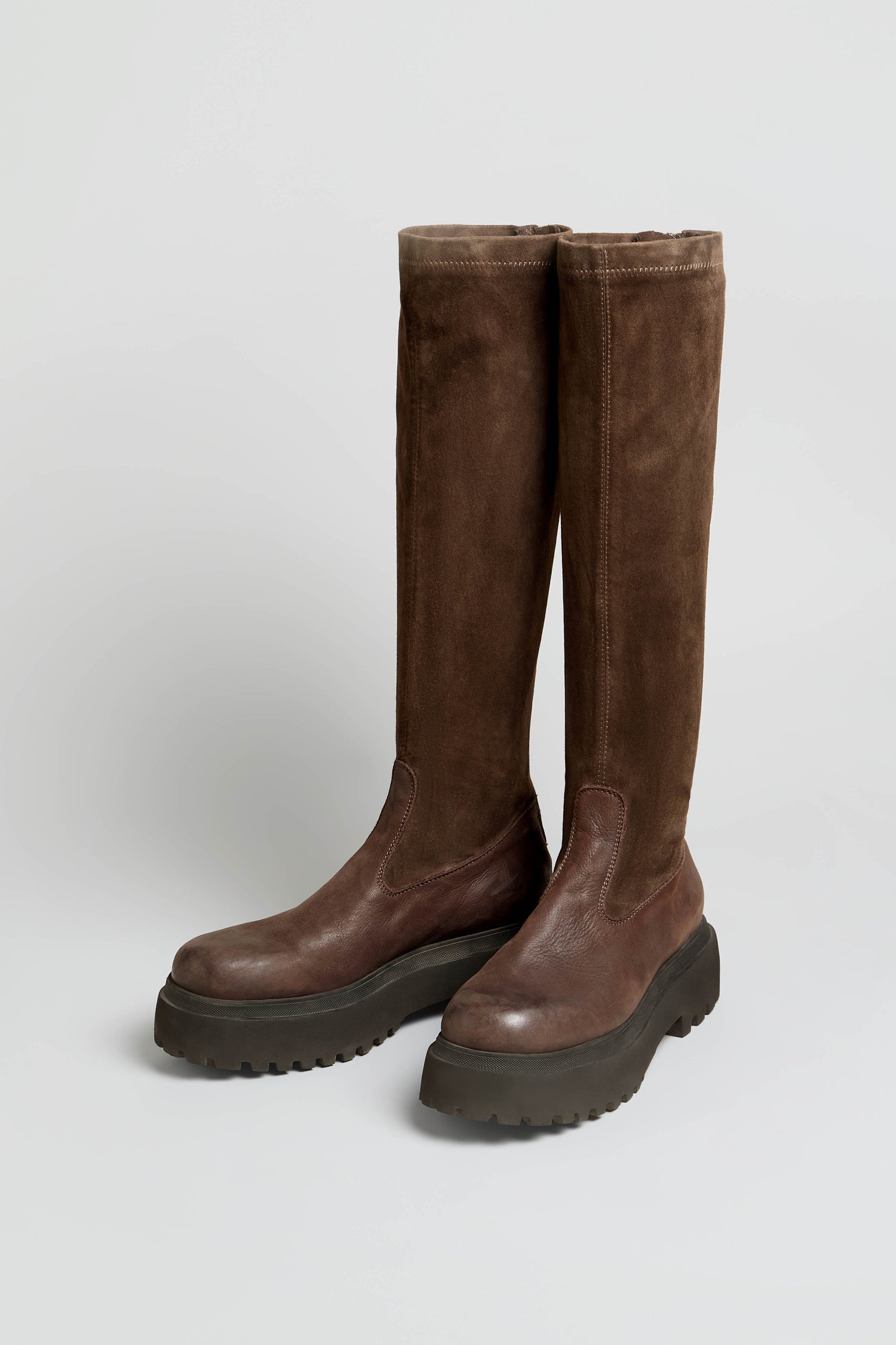 A pair of Knee High Stretch Suede Boots in a rich brown shade, featuring thick black rubber soles and rounded toes, positioned side by side on a plain light background.