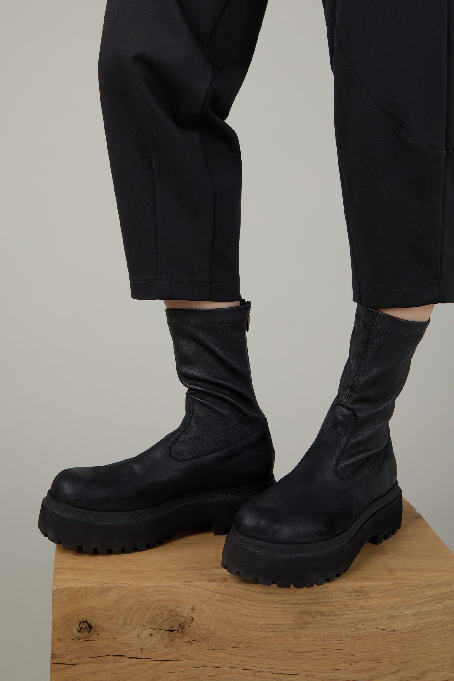 Someone wearing Short Stretch Leather Black Boots with a chunky platform sole and black pants stands on a wooden platform against a plain background.