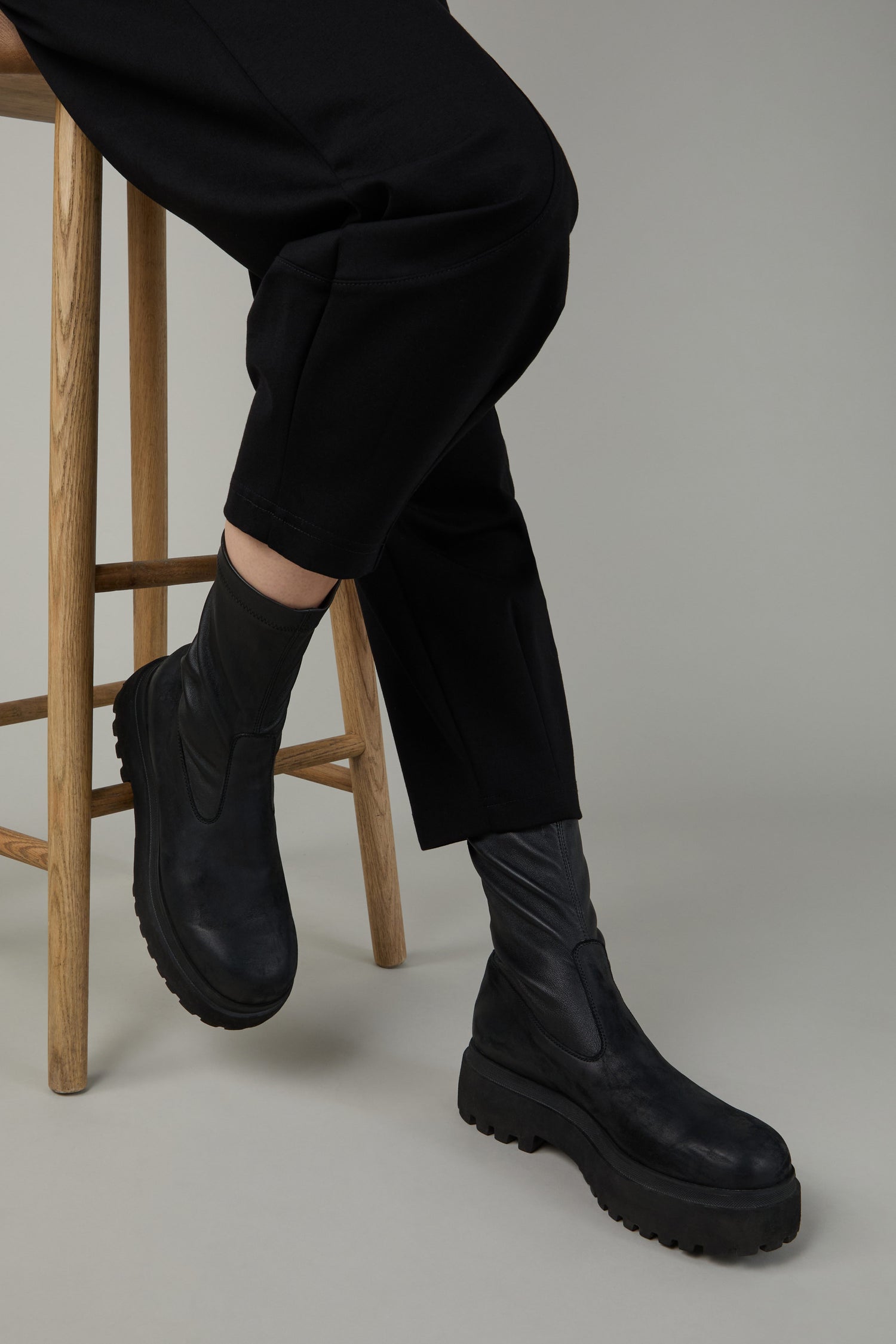 Wearing black pants and Short Stretch Leather Black Boots, a person with platform soles sits on a wooden stool against a plain background.